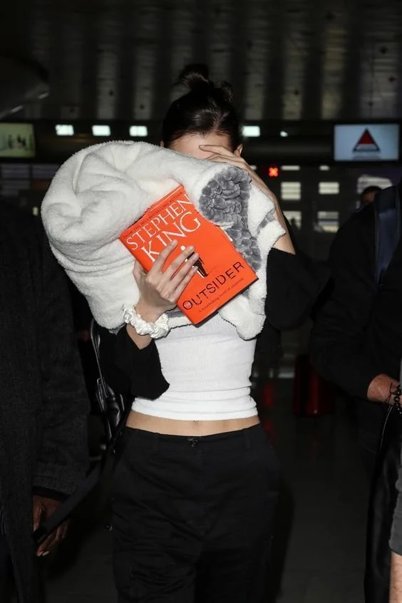 Kaia Gerber holding a copy of Stephen King’s The Outsider in front of her face while walking indoors.