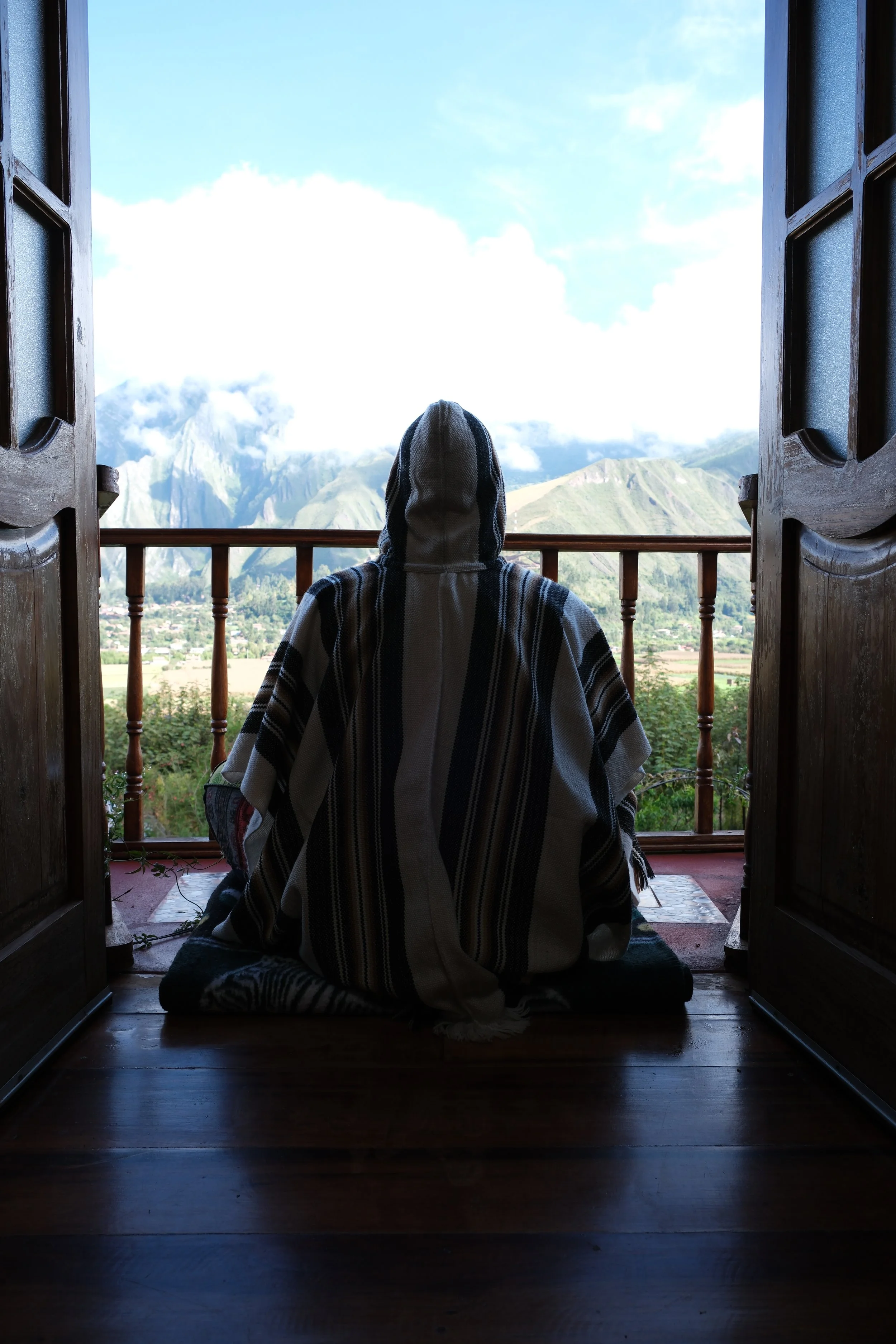 Person sitting on a wooden floor, facing a balcony with mountains in the distance, wearing a hooded blanket, viewed from behind.