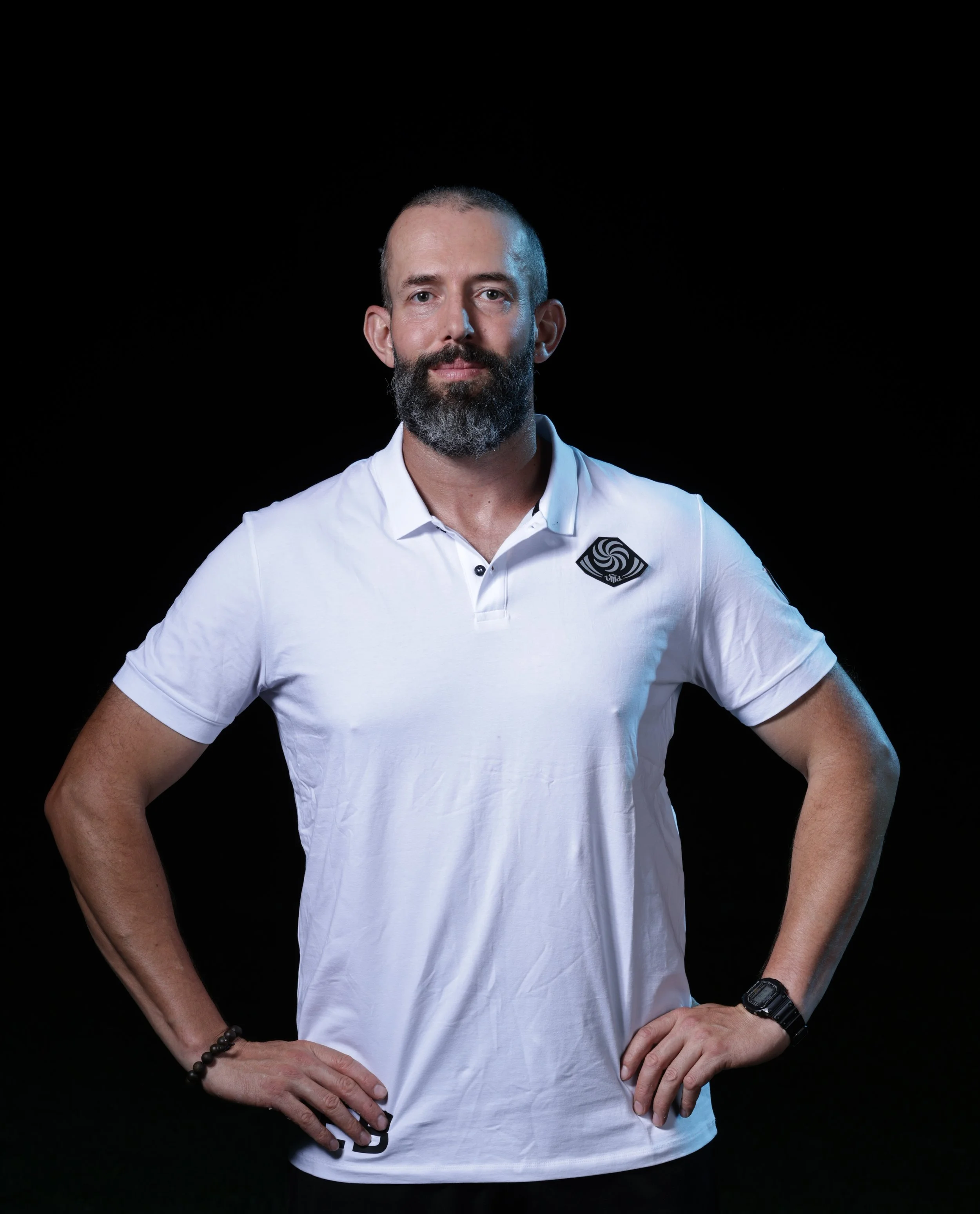 A man with a beard and shaved head, wearing a white polo shirt with a black and white logo on the chest, standing against a black background with his hands on his hips, looking directly at the camera.