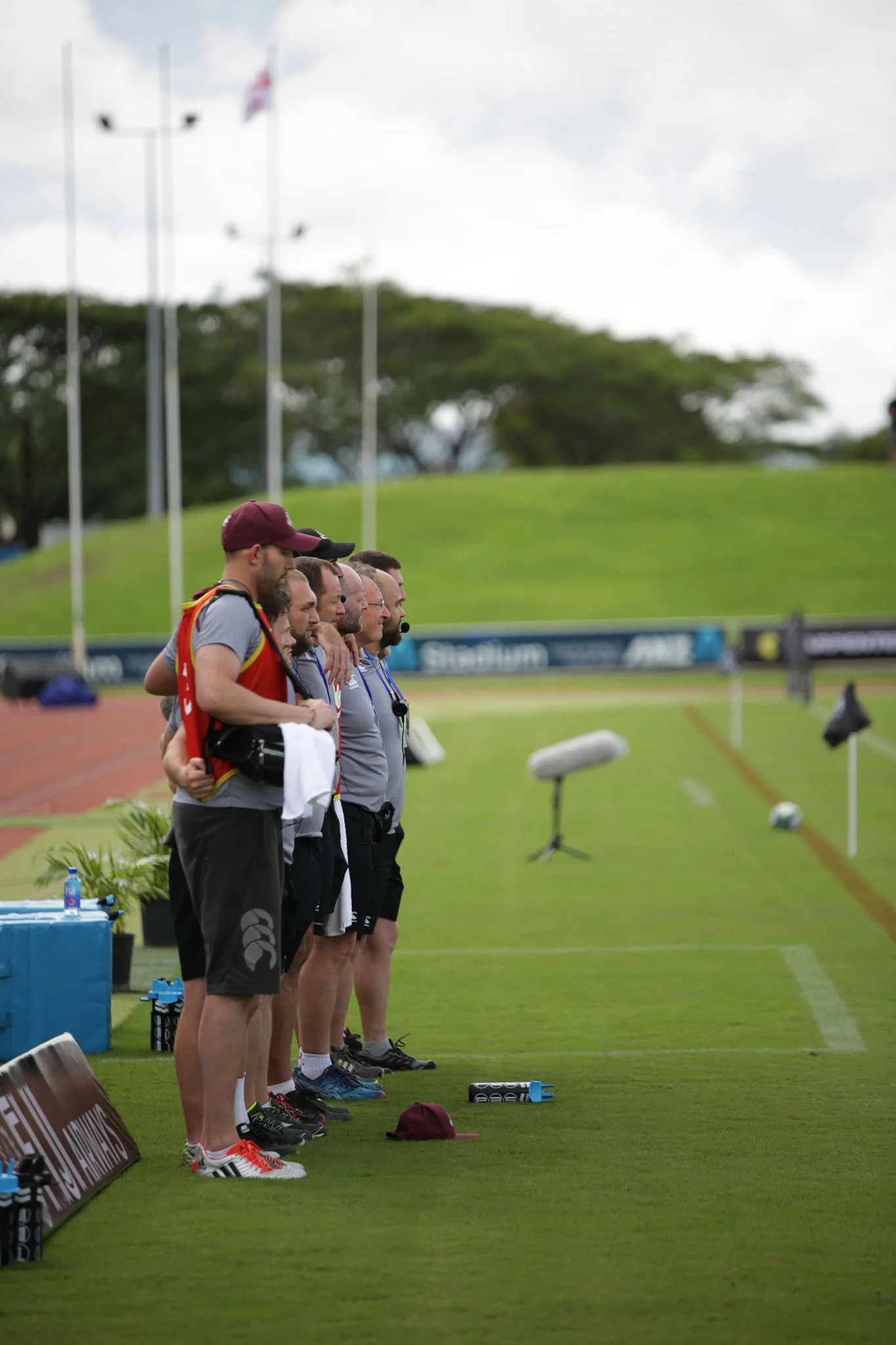 Group of people on the sidelines of a sports field, standing in a row and watching a game, with flags and trees in the background.