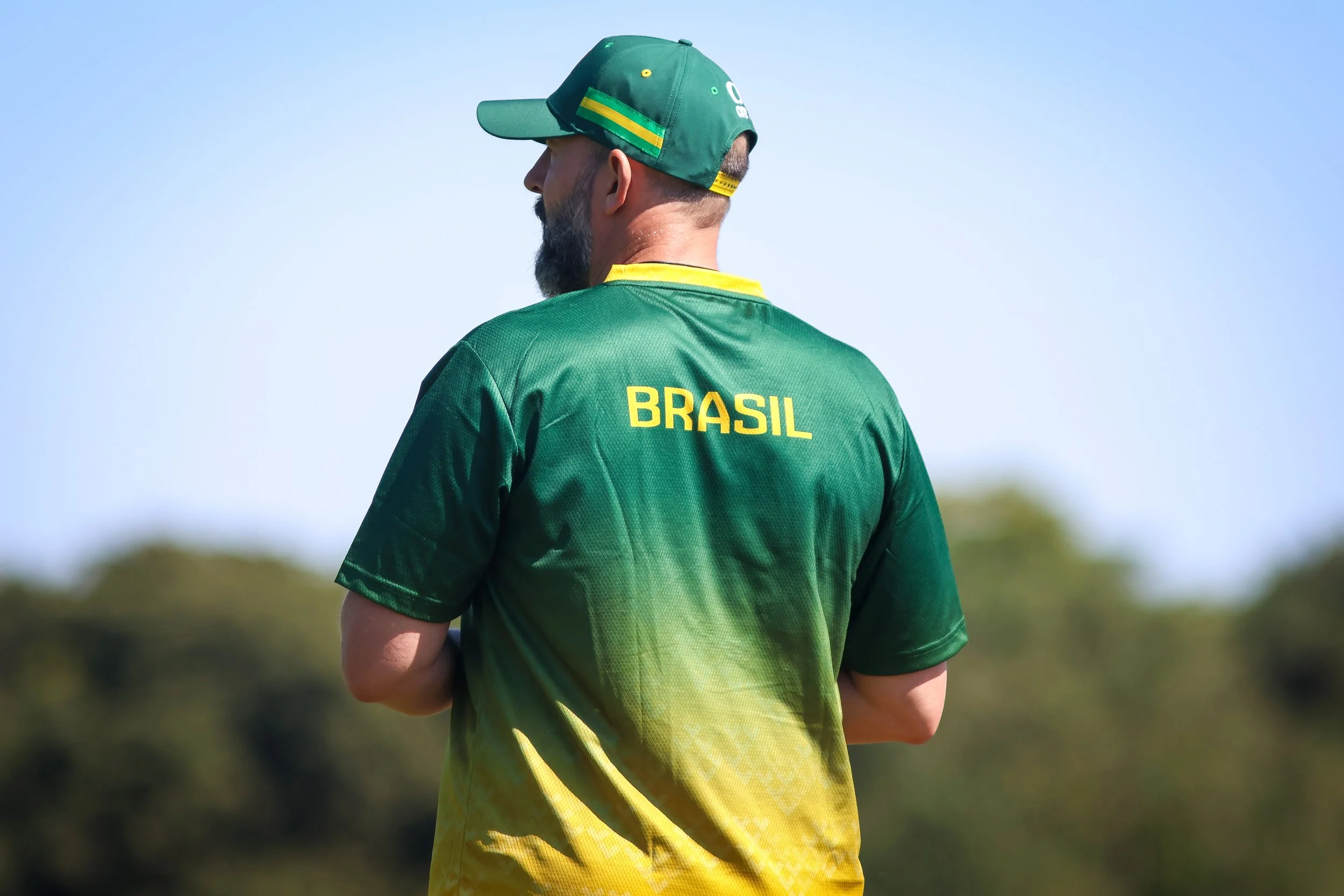 A man wearing a green and yellow Brazil national football team jersey and a green cap. He is standing outdoors with a clear blue sky and blurred trees in the background, facing away from the camera.
