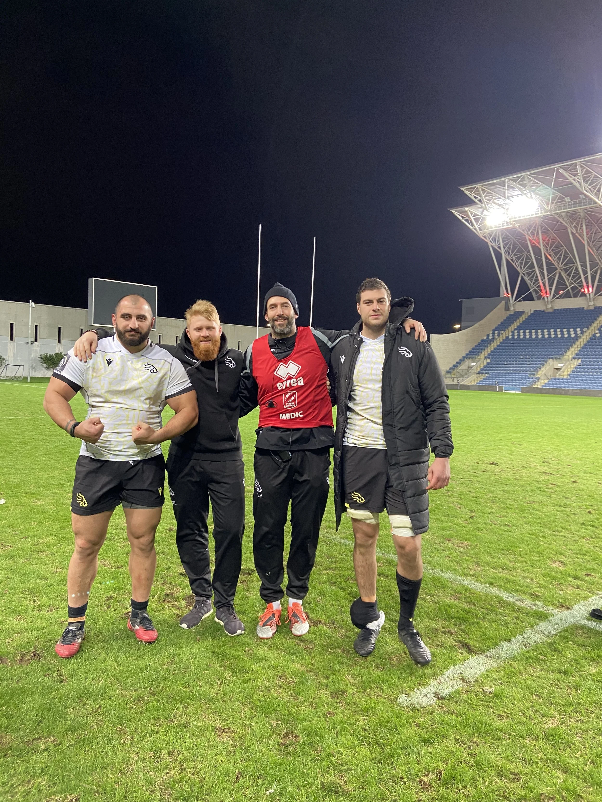 Four rugby players standing on a field under stadium lights at night, smiling and posing with their arms around each other, one wearing a red training vest.