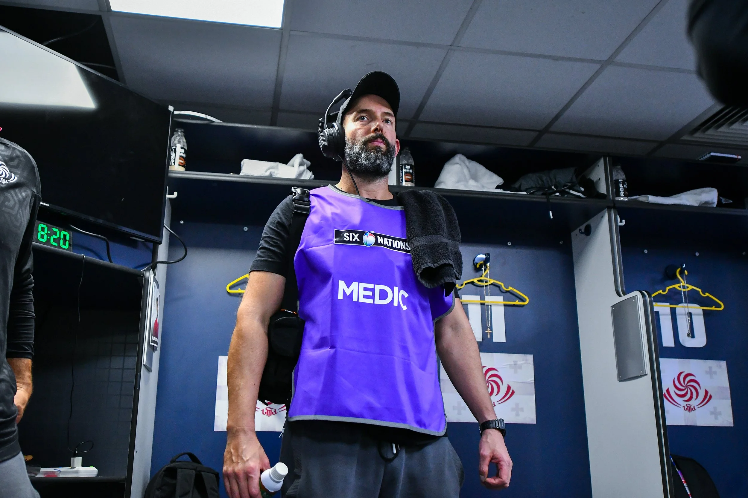A man with a beard wearing a blue medical vest and black cap, standing in a locker room with a towel over his shoulder, holding a water bottle, and listening to headphones.