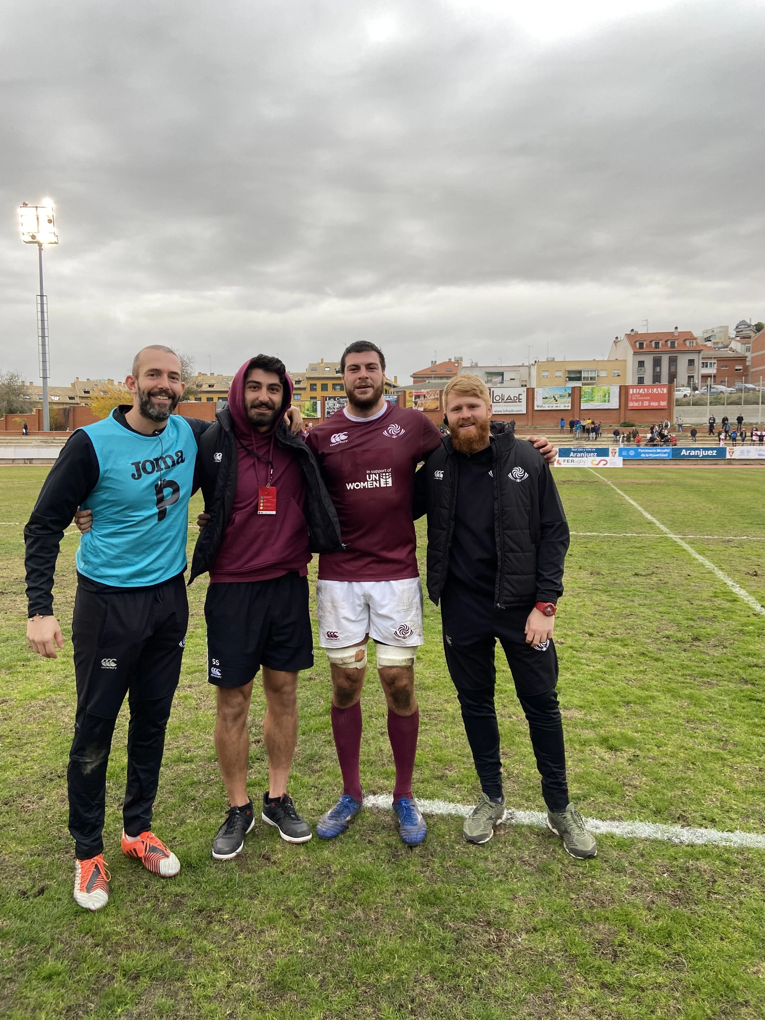 Four men standing on a rugby field with their arms around each other's shoulders, smiling. Three are wearing sports clothing, and one is in a black jacket. Overcast sky, sports stadium in the background.