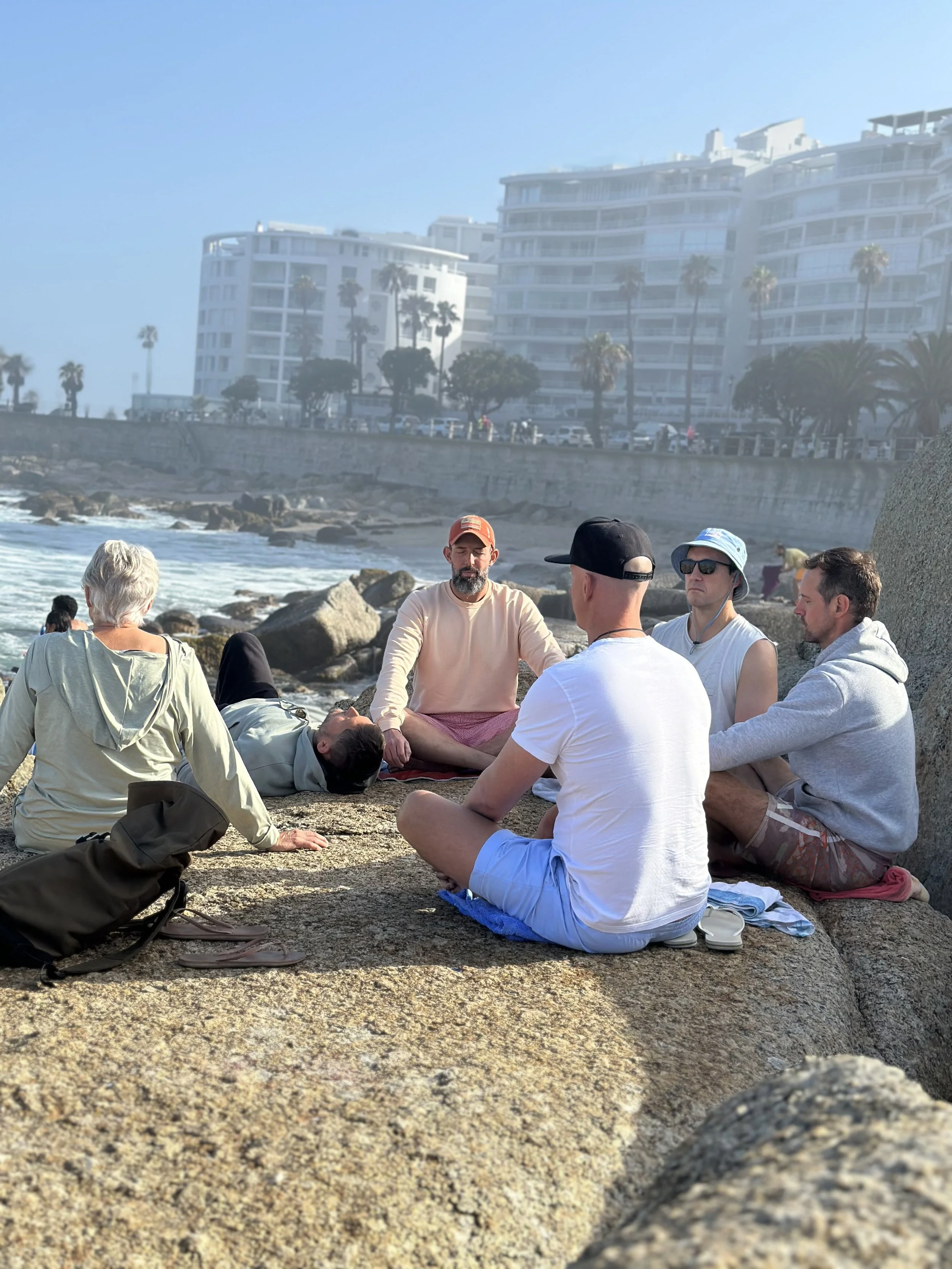 Group of six people practicing meditation or yoga on rocks by the beach with buildings and palm trees in the background.