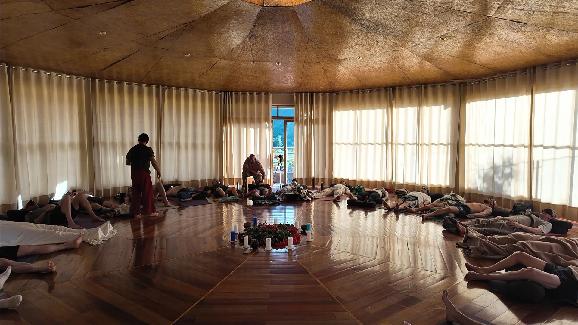 A group of people practicing yoga or meditation in a circular room with large windows and wooden floors. Some are lying down, some are sitting, and there are candles and flowers arranged in the center.