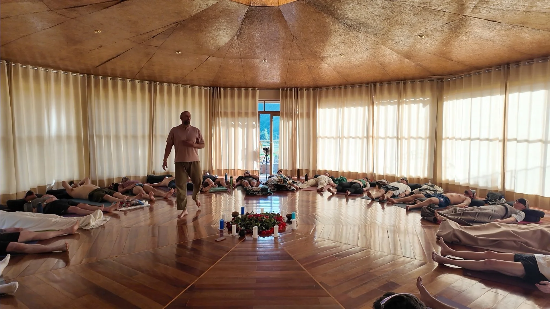 A group of people lying on the wooden floor of a circular room with cream curtains and a man walking among them, possibly during a meditation or yoga class.