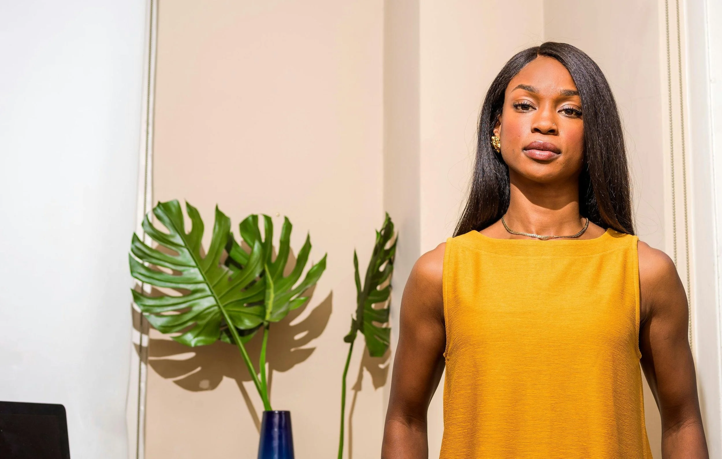 A black woman with long straight hair and a sleeveless yellow top standing in front of a desk.
