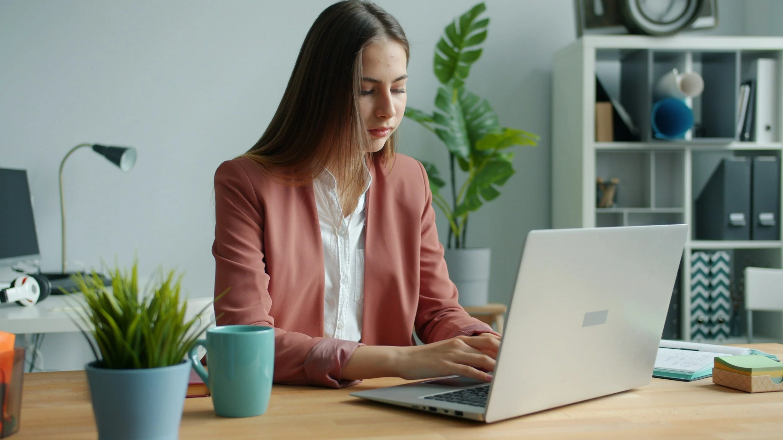 A young white woman sitting at a desk typing onto a laptop, wearing a salmon pink blazer.