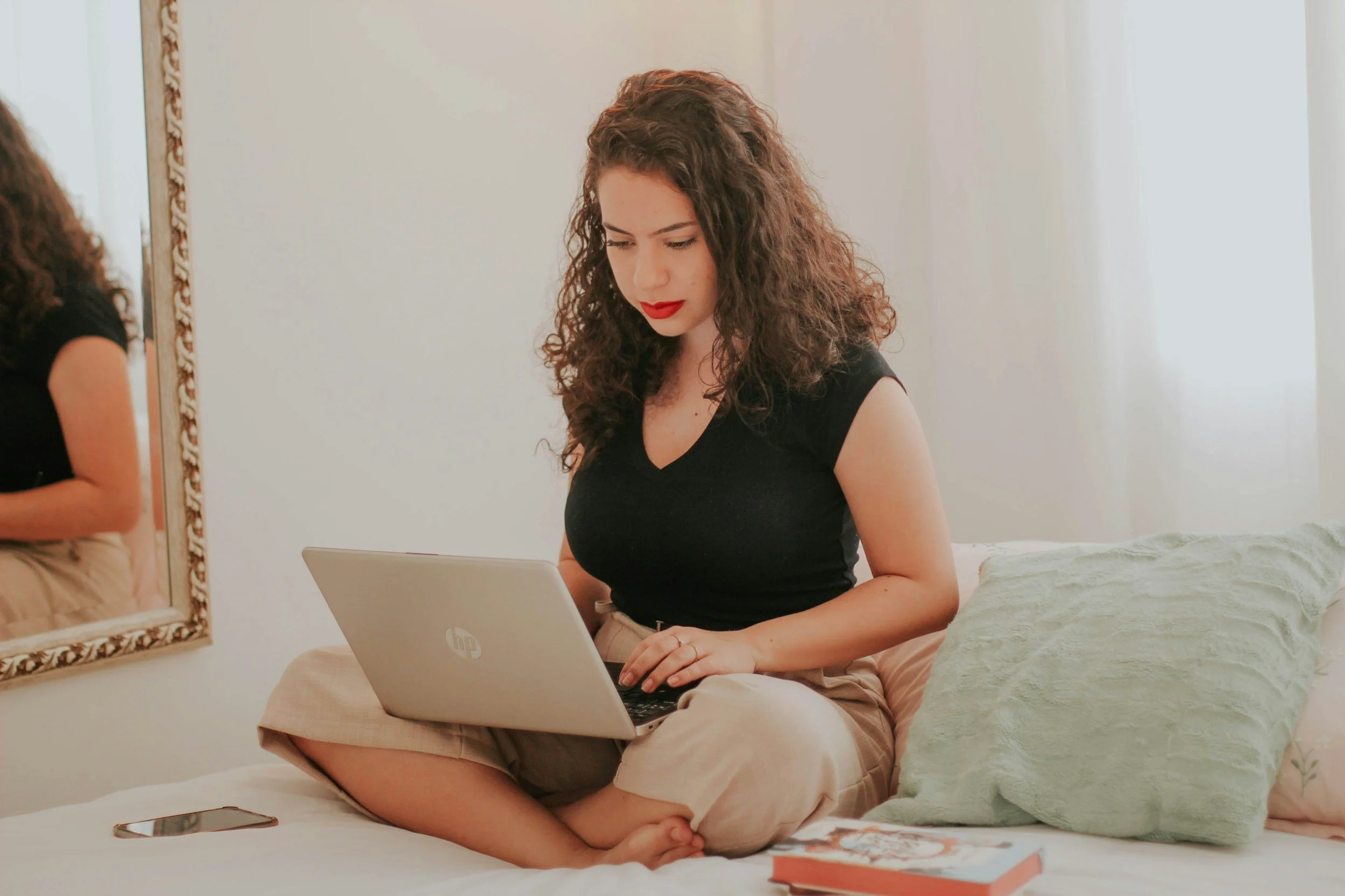 A racially-ambiguous woman on a laptop in bed beside a mirror.