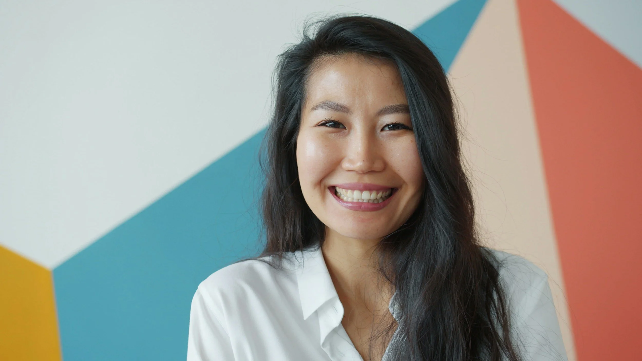 Asian woman smiling in a white blouse with long black hair against a multicoloured wall.