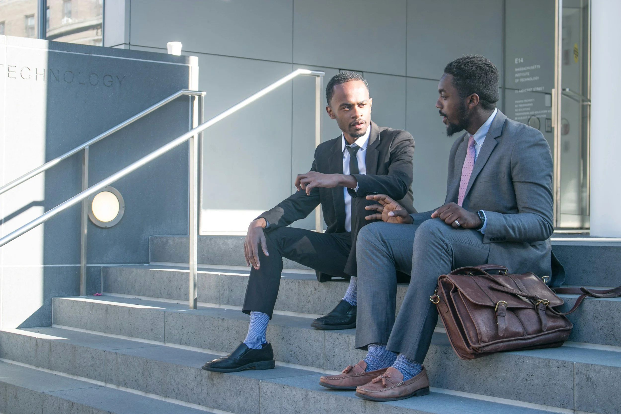 Two black men in suits sitting on steps talking.
