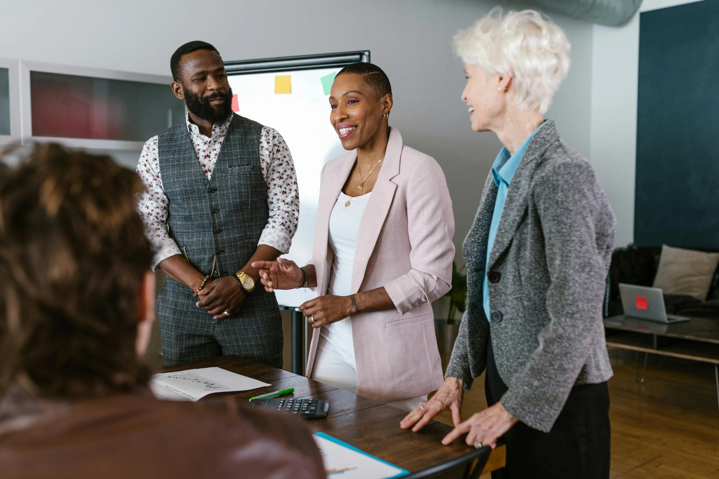 A black man, a black woman and a white woman smiling at a room full of people in a boardroom.