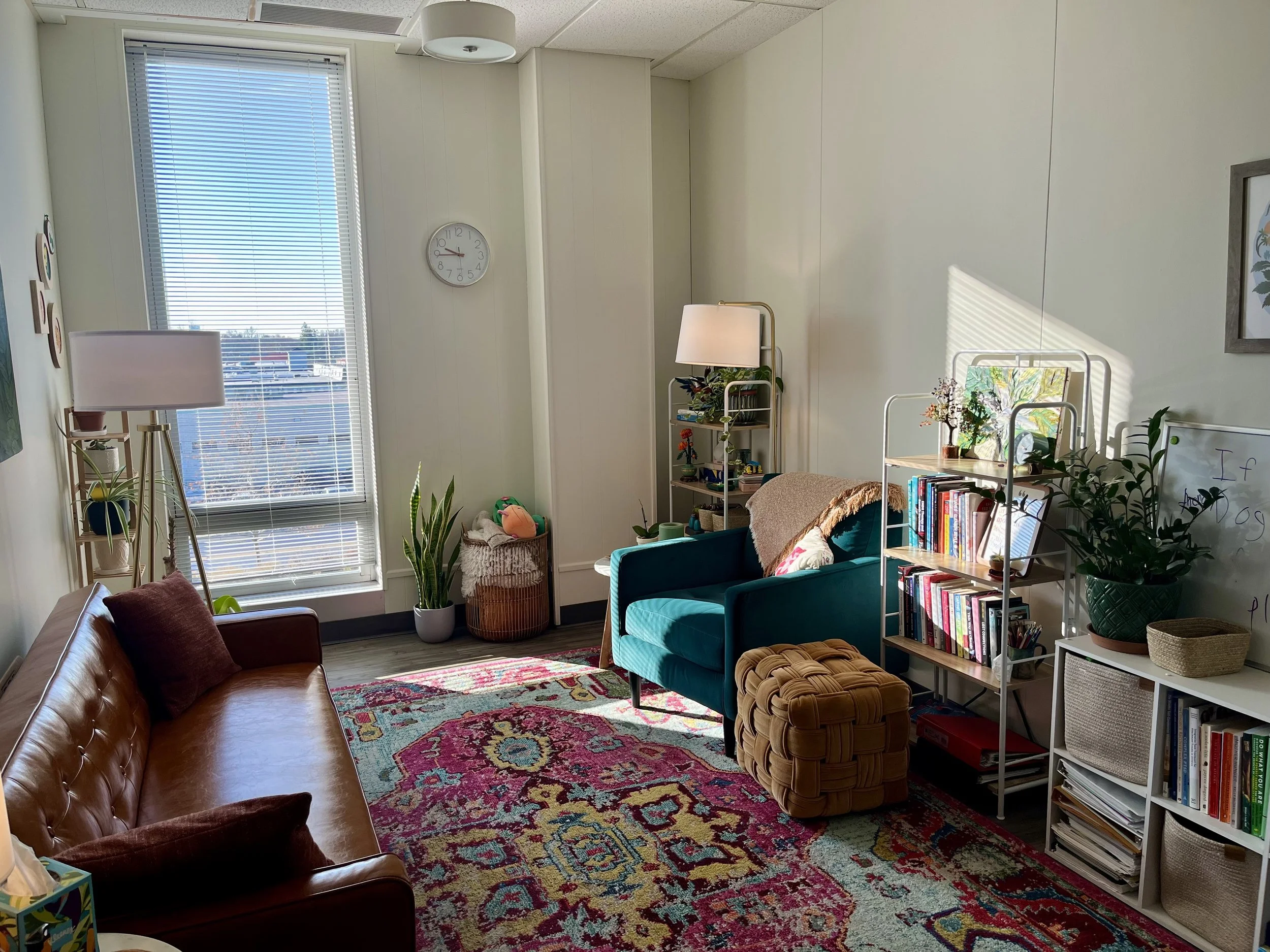 A cozy therapy office with a large window letting in sunlight, a brown leather couch with pillows, a teal armchair with a blanket, two white bookshelves filled with books and plants, a colorful patterned rug, and various decorative items.