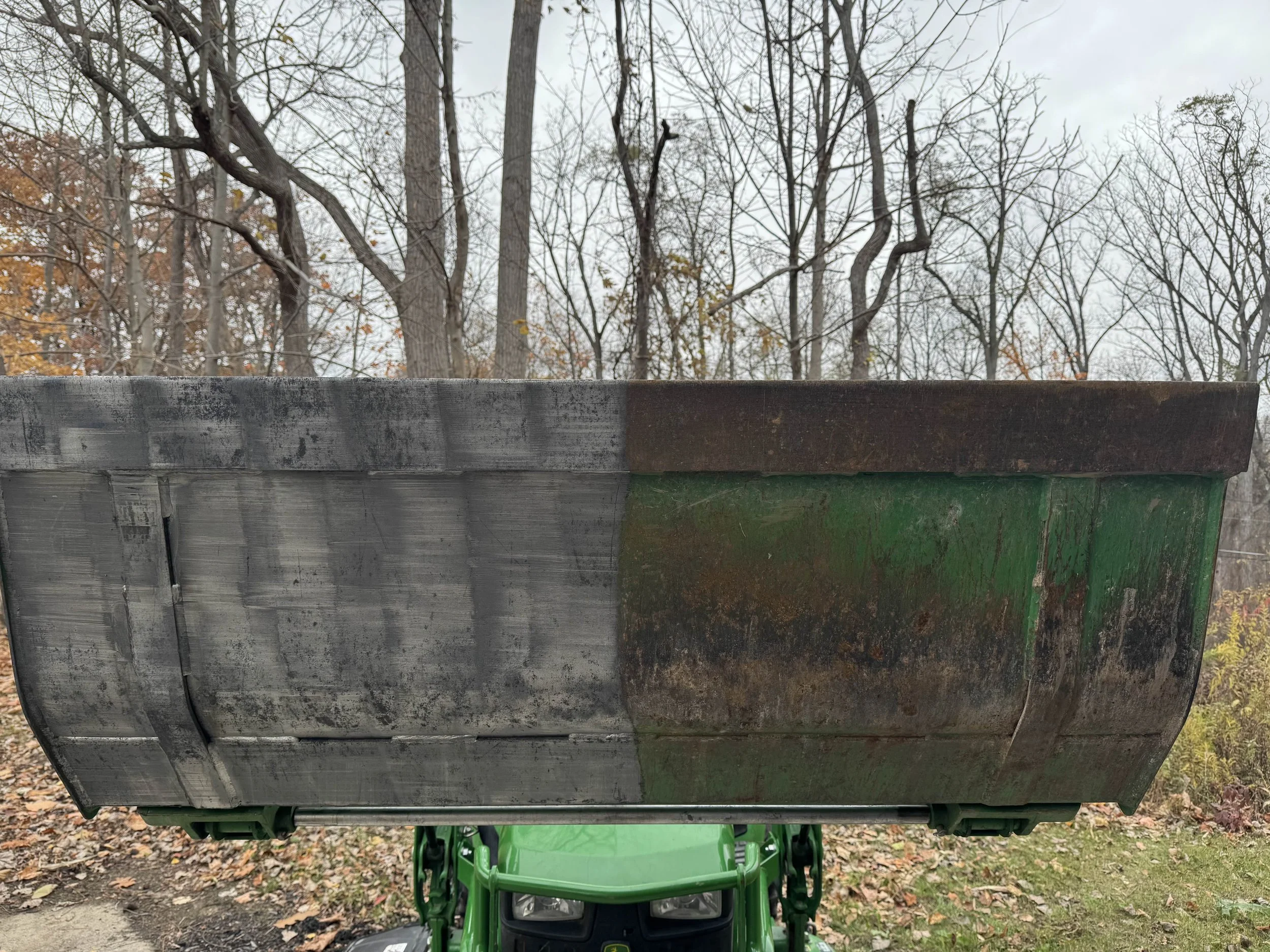Close-up of a green tractor with a large rusty metal scoop or bucket attached, set outdoors with leafless trees and autumn foliage in the background.
