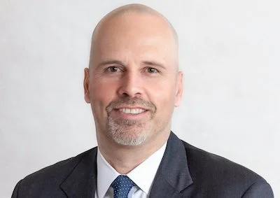 A headshot of a bald man with a goatee, wearing a dark suit and tie, smiling against a light gray background.
