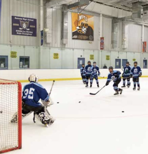 Young hockey players practicing shooting pucks at an indoor ice rink, with a goalie in blue gear and multiple players in blue jerseys and helmets.