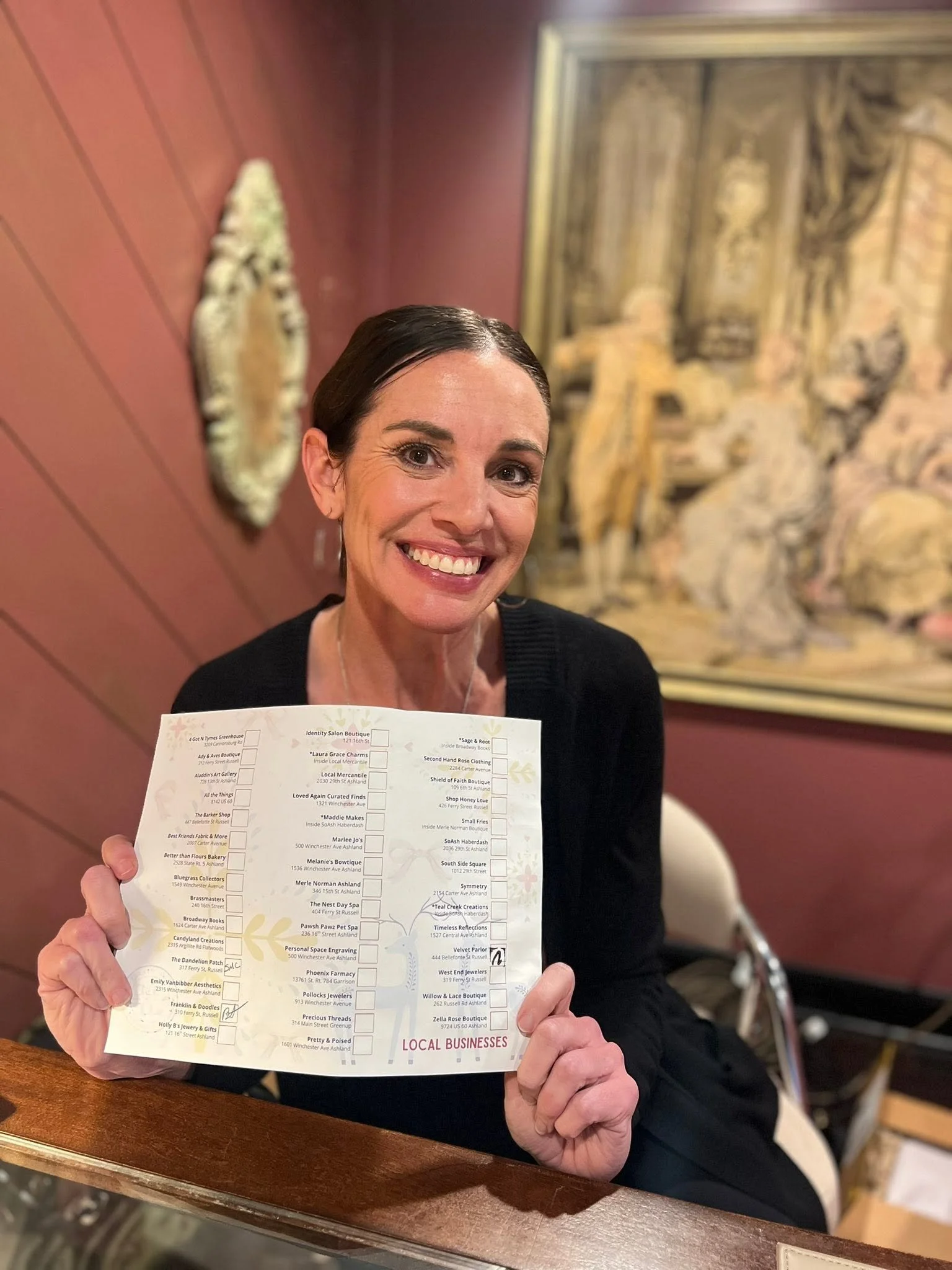 A woman with dark hair and a big smile is showing a printed list of local businesses, holding it up with both hands. The background features a large ornate mirror and a red wall.