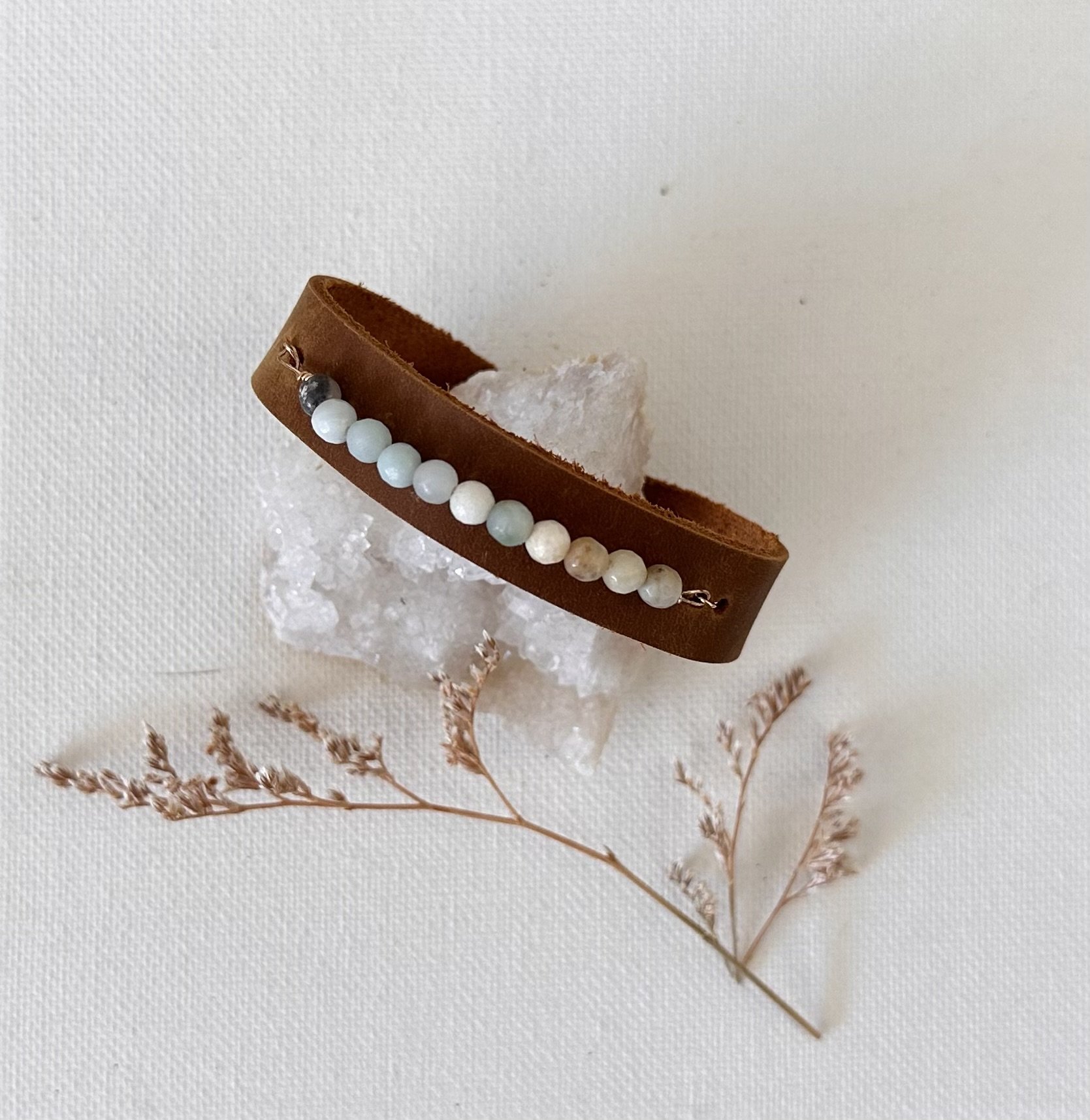 Leather bracelet with white beads, resting on a crystal, with dried pinkish-brown twigs underneath, all on a white surface.