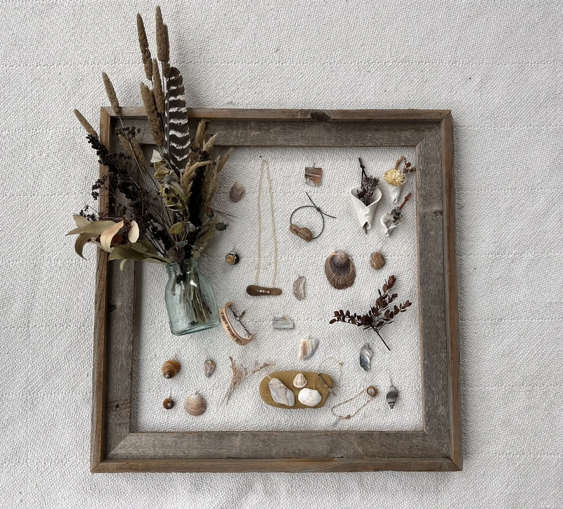 A wooden frame on a beige textured background contains an arrangement of dried plants, shells, and jewelry pieces. The dried plants include feathers and grasses in a small glass jar, and there are various shells and small decorative items scattered inside the frame.