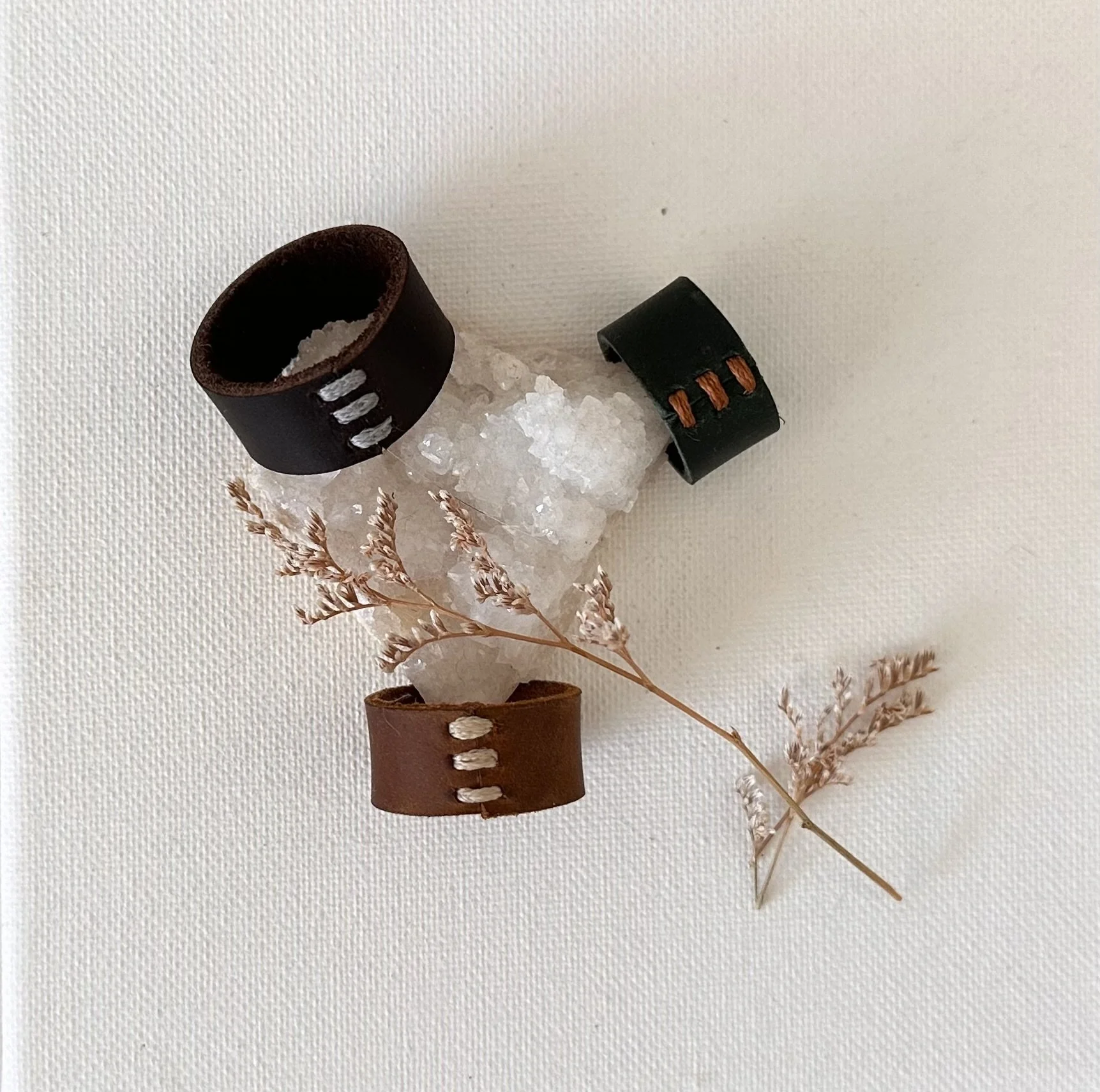 Leather wristbands, salt crystal, dried pinkish-brown sprig on white textured background.