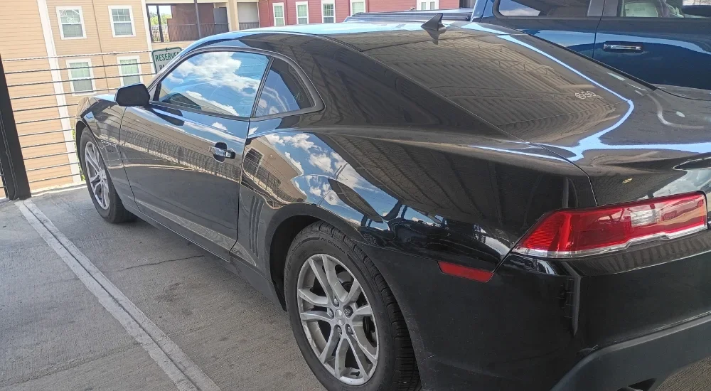 Black coupe car parked in a parking lot, with buildings and a reflection of the sky on the car's surface.