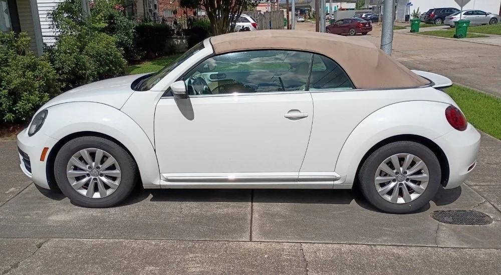 A white Volkswagen Beetle convertible with a tan soft top parked on a sidewalk.