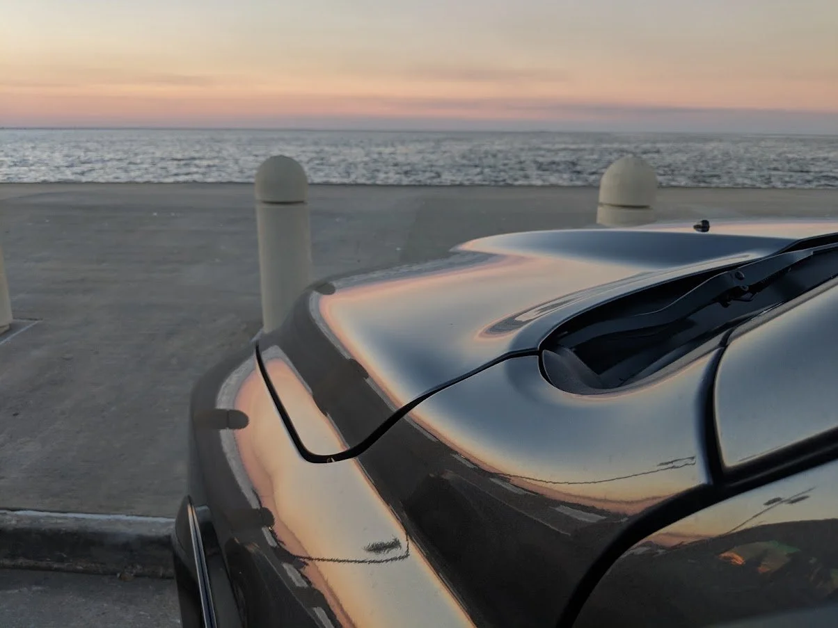 Close-up of a black car's roof and windshield reflecting a colorful sunset over Lake Pontchartrain.