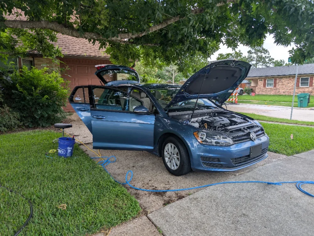 A blue Volkswagen car with the hood, driver's side door, and rear hatch open, parked on a driveway. It appears to be in repair or maintenance mode, with a sponge, bucket, and hoses nearby. The scene is outdoors in a residential neighborhood.