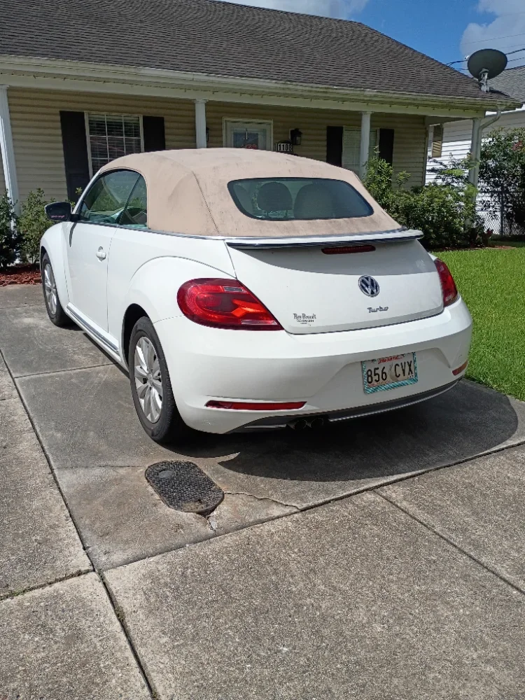 White Volkswagen convertible with a beige soft top parked on a concrete driveway in front of a yellow house with black shutters and green lawn.