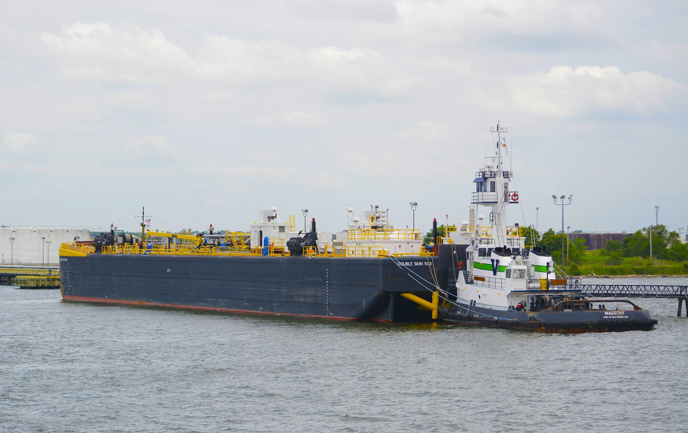Oil tanker docked at a port with cloudy sky, trees, and infrastructure in the background.