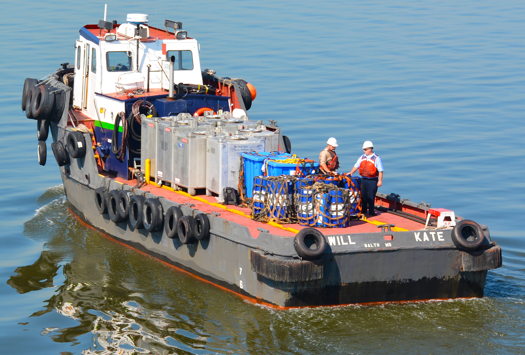 A tugboat named 'Will Kate' sailing on calm water, with two crew members wearing safety helmets and vests, surrounded by large containers, hoses, and equipment on its deck.