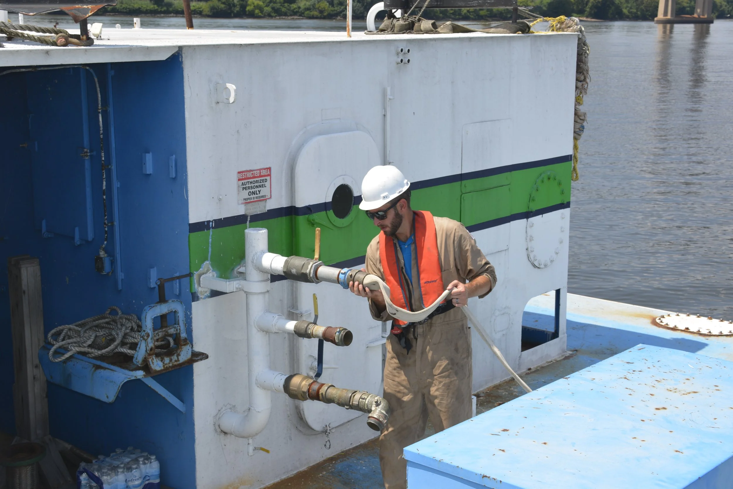 A worker in safety gear, including a white helmet and sunglasses, handling a hose on a boat or ship near water, with pipes and equipment visible on the vessel.