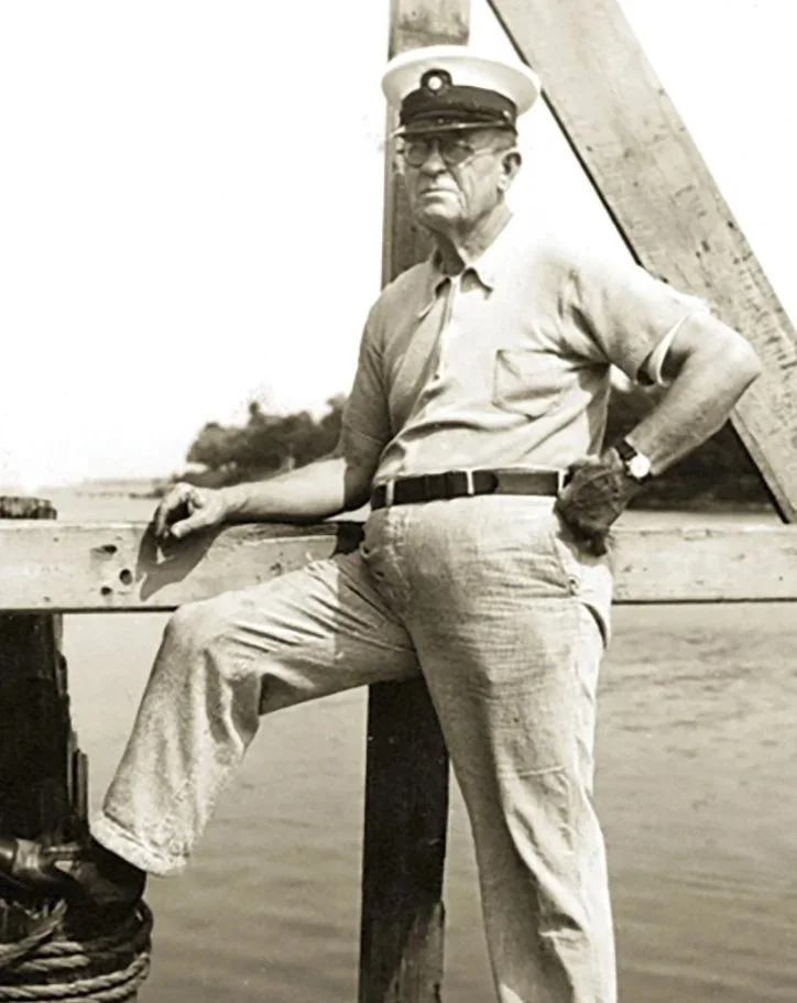 A black and white photo of a man in a sailor or captain's cap, glasses, light-colored shirt, and light-colored pants, standing on a boat or dock with one foot resting on a ledge and one hand on his waist, with water and a distant shoreline in the background.