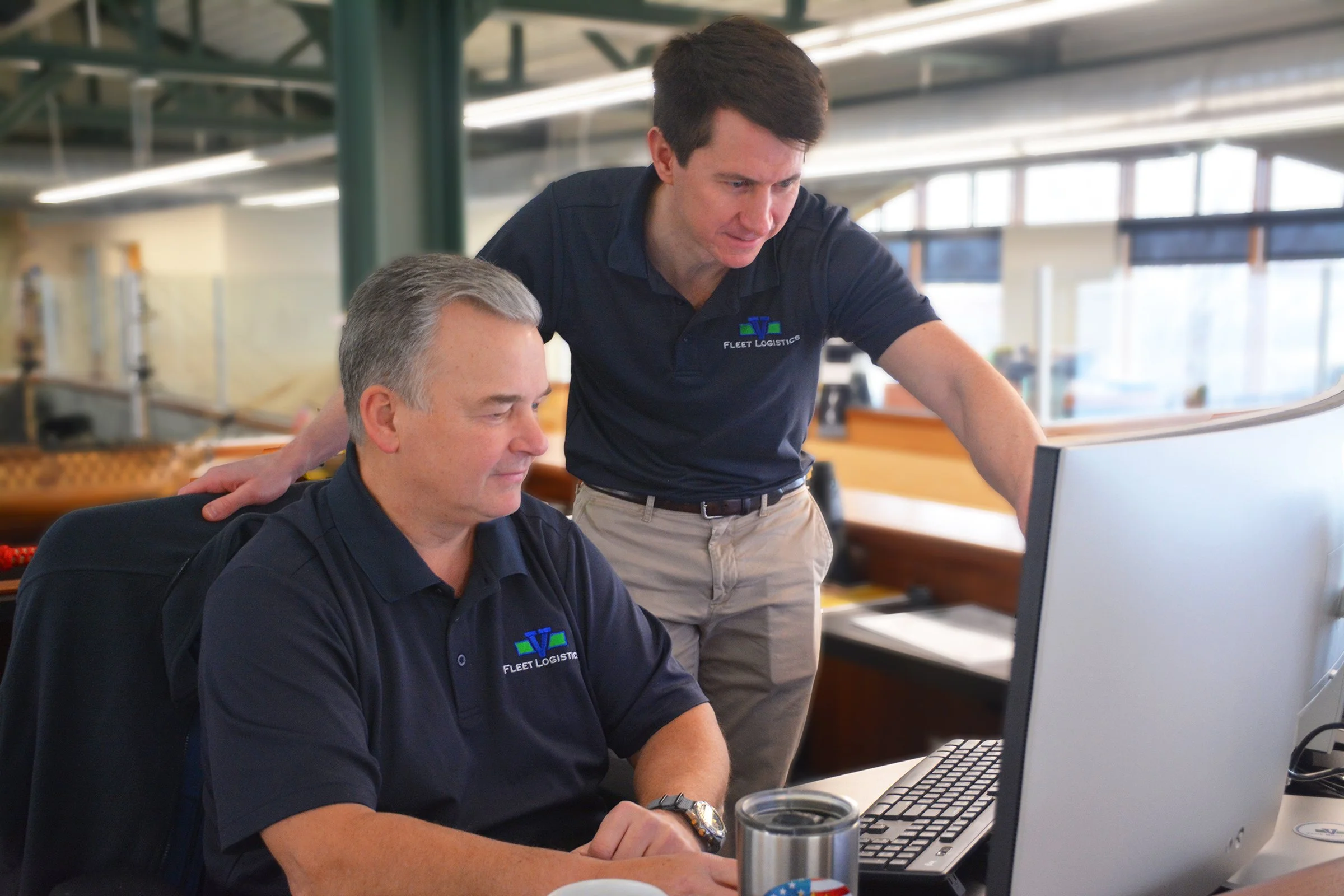 Two men wearing navy blue shirts with the Fleet Logistics logo working together at a computer in an office setting.