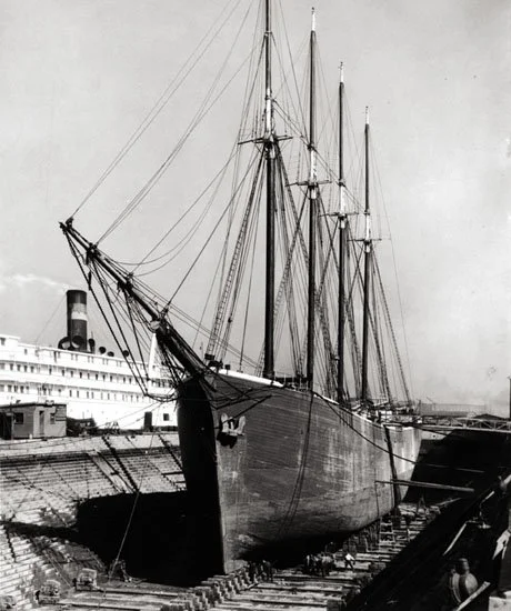 Black and white photo of a large sailing ship docked in a shipyard with a cityscape in the background.
