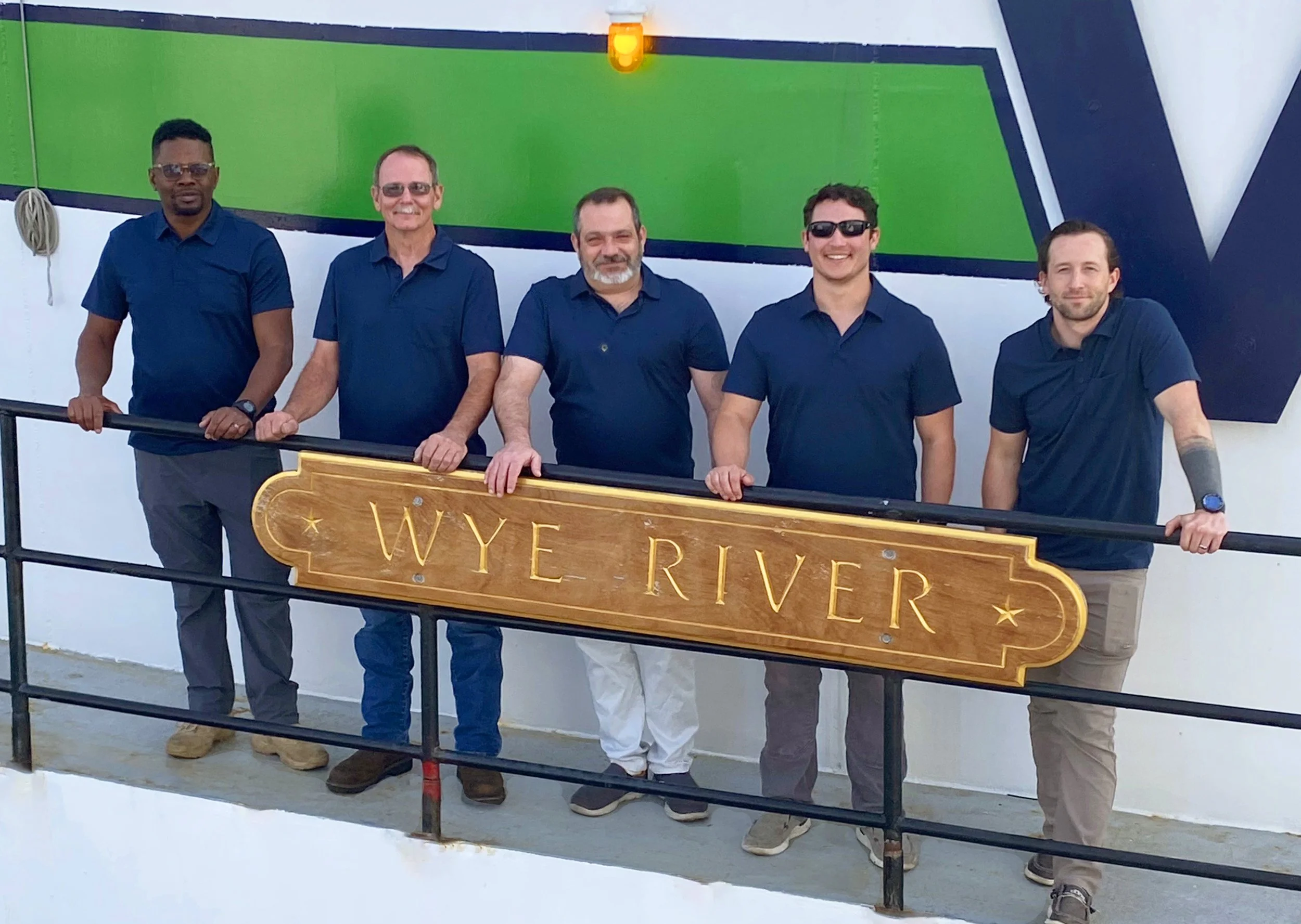 Five men standing behind a wooden sign that reads "WYE RIVER," all wearing dark blue shirts, some with sunglasses, smiling, against a background with green, black, and white colors.