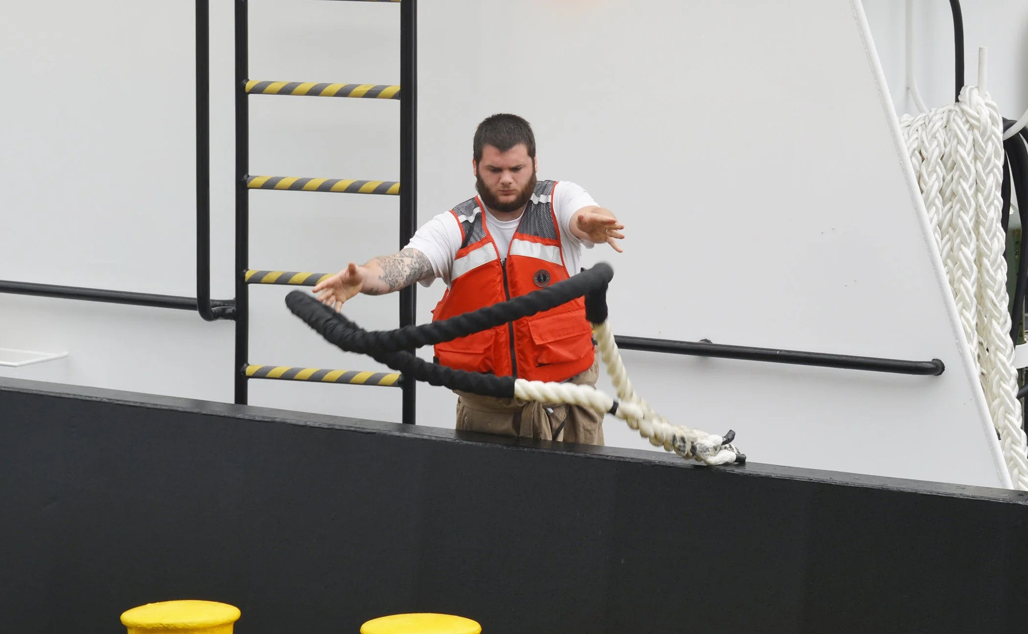 A man with a beard and tattoos on his arms wearing an orange safety vest over a white shirt and tan pants is pulling on a thick rope with a loop, standing on the deck of a ship or boat.