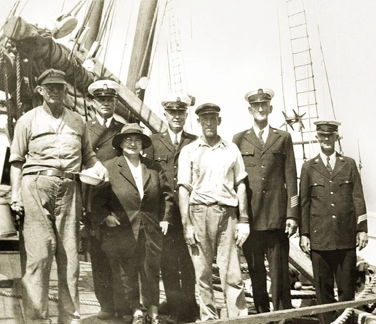Seven people, including men in military uniforms and other attire, standing on a ship's deck with rigging and ropes visible in the background.