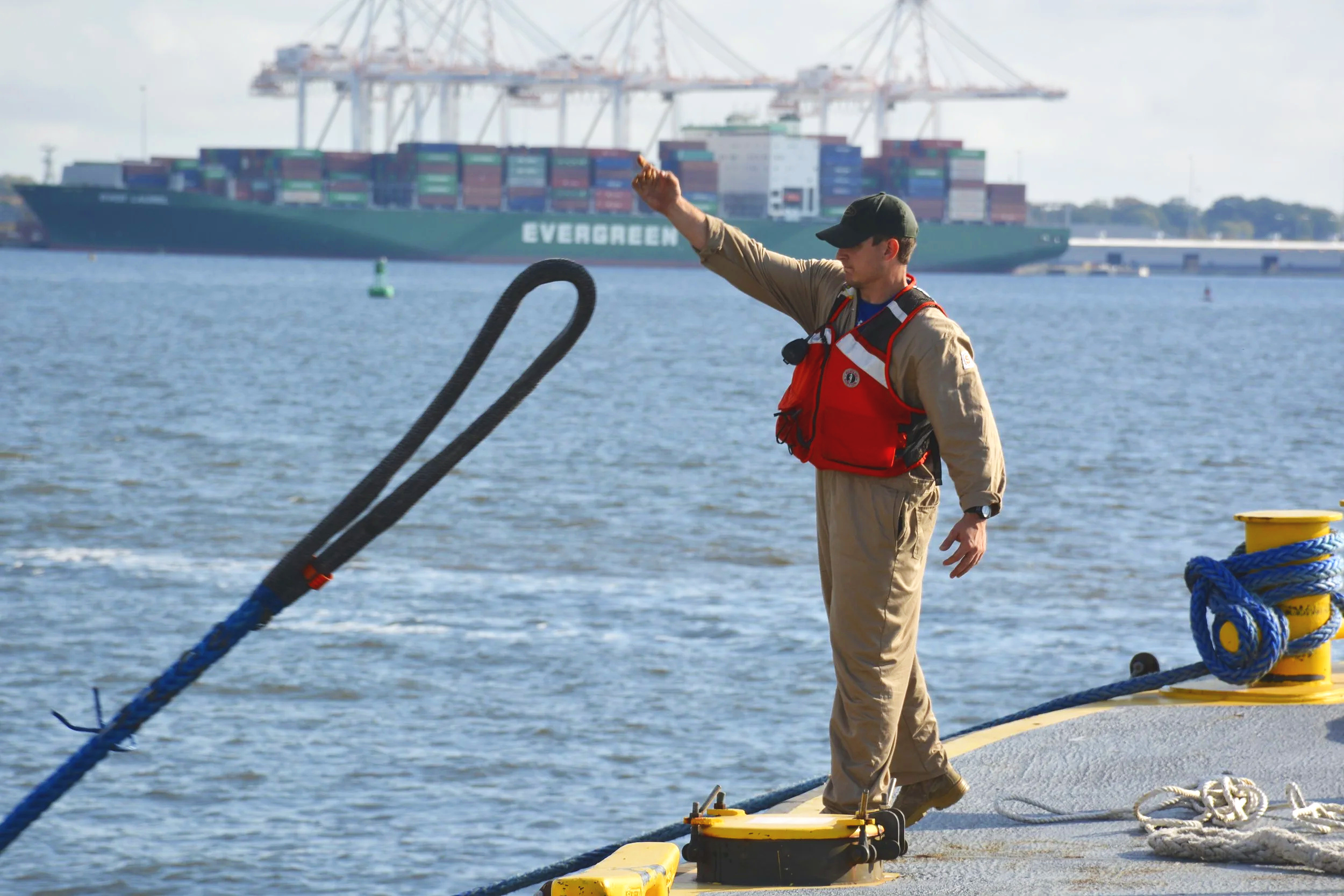 A person in uniform and a red life jacket standing on a dock, holding a large black tow line in the water with a container at their feet, with a cargo ship in the background.