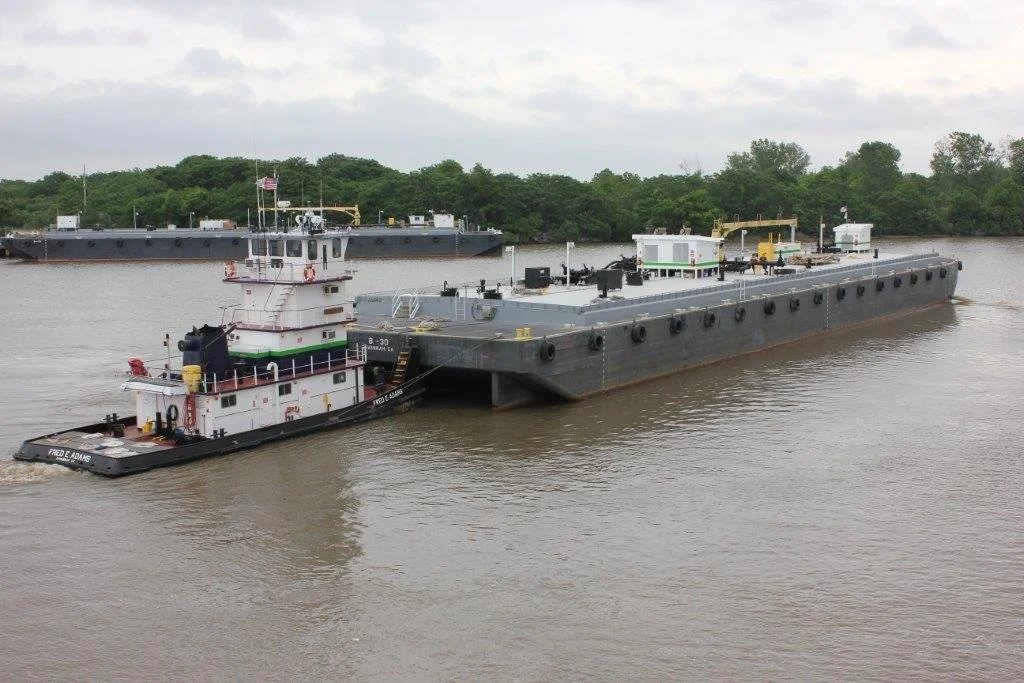 A large gray barge being pushed by a tugboat on a river, with another barge in the background and green trees along the riverbank.