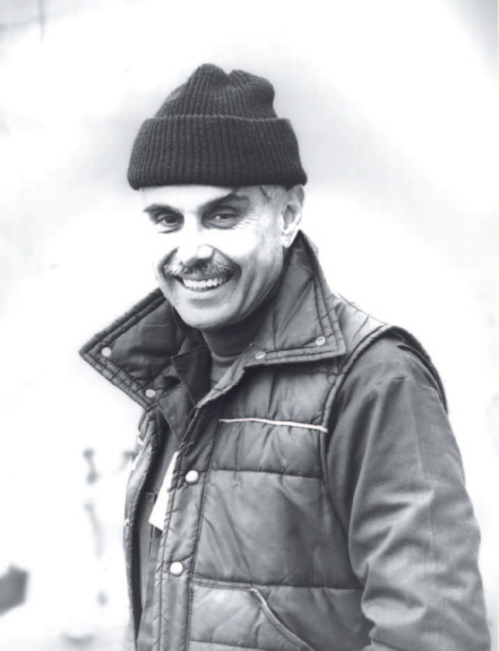 A middle-aged man with a mustache, wearing a knit beanie, a puffy jacket, and smiling outdoors.