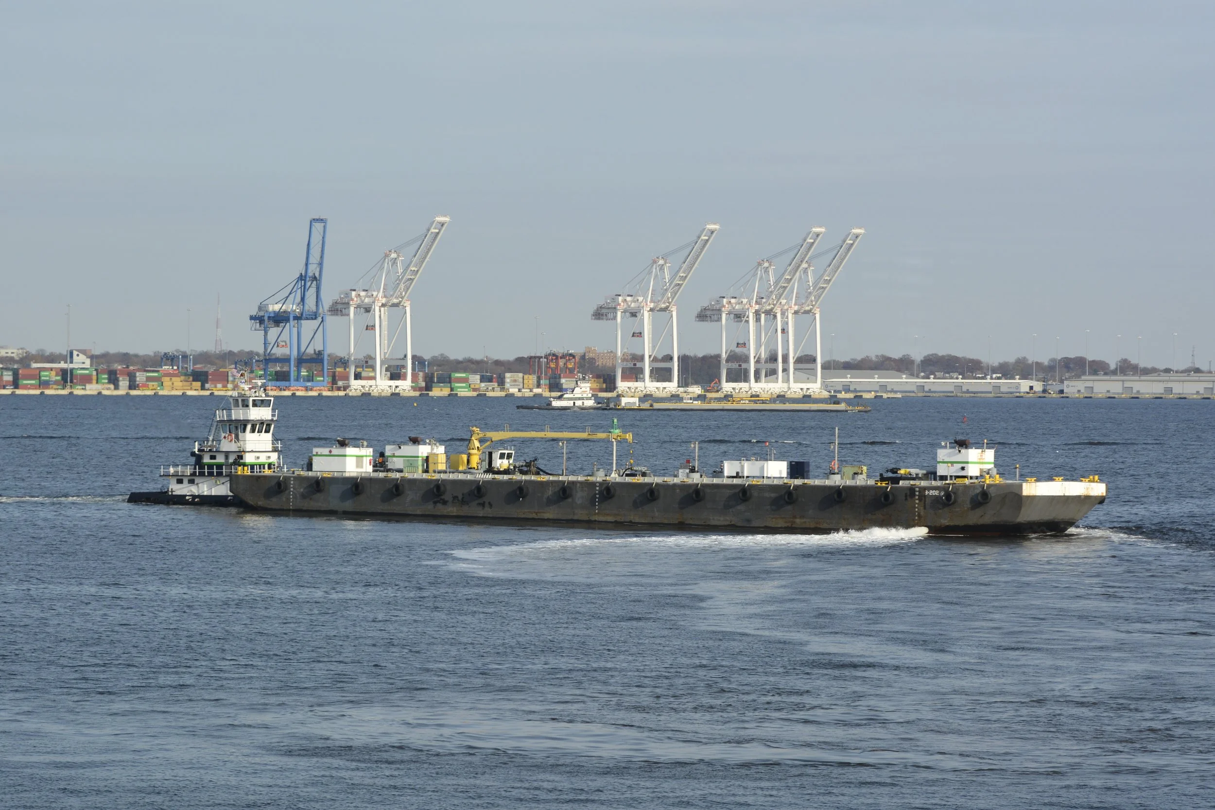 A cargo ship sailing on the water with container cranes and stacked containers in the background.