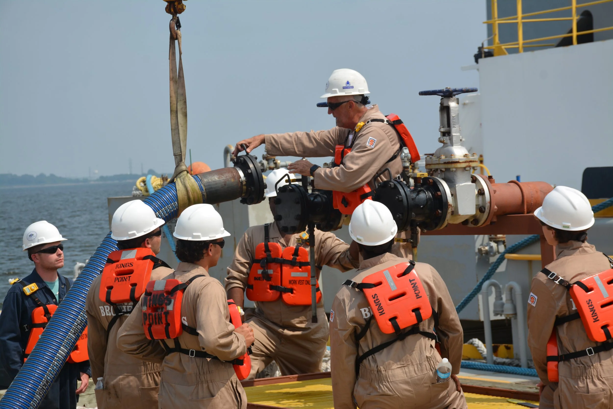 A group of workers in safety gear, including white helmets and orange life vests, surrounding a worker operating a control valve on a large industrial pipe on a vessel at sea.