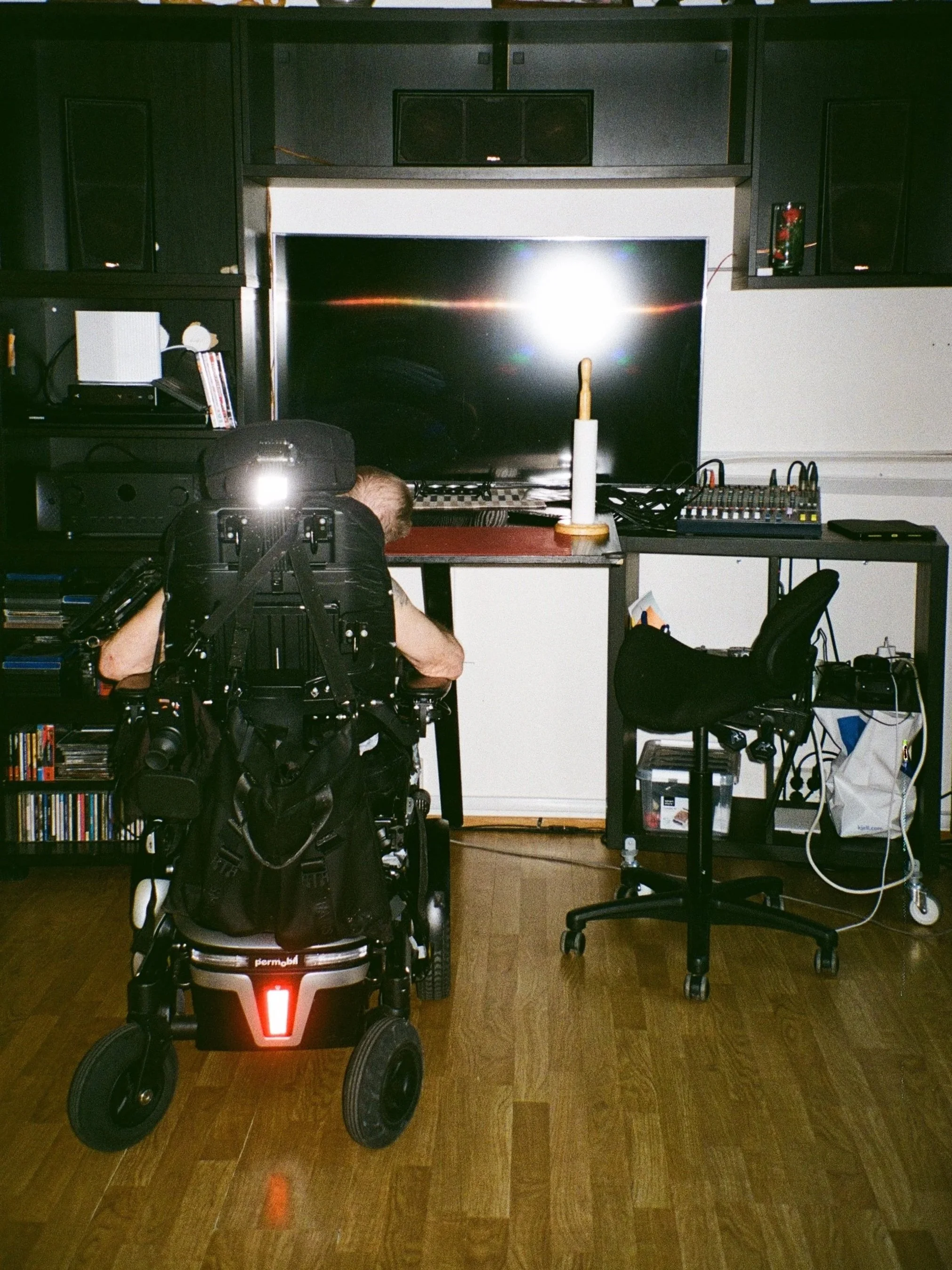 foto av svart møblement langs vegg. hyller rundt og over med en tv i midten. bordet stikker ut fra tv. en person sitter i rullestol foran bordet og tvn.