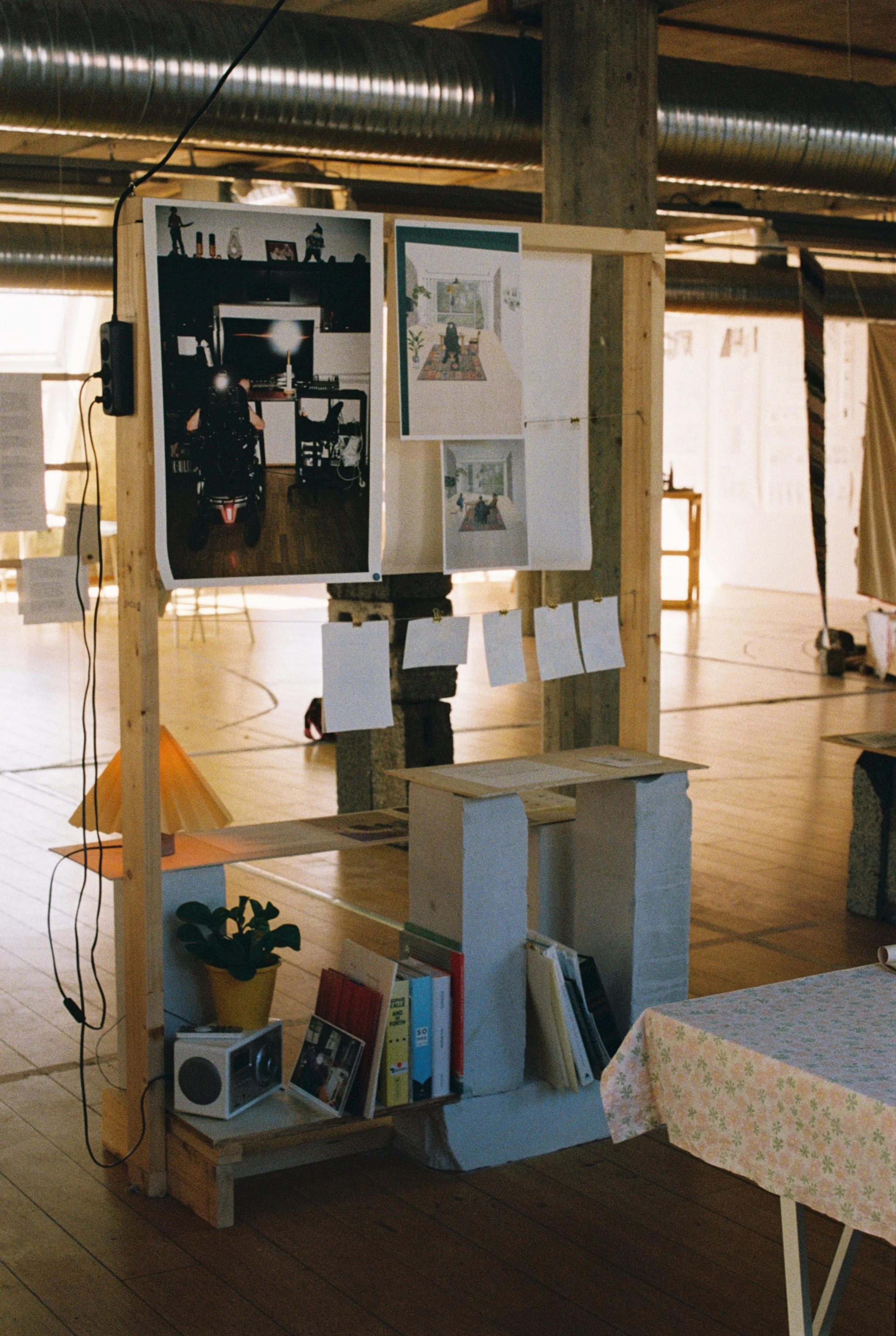 Foto fra utstilling. stativ med hyller. Stativet har store fotografier og illustrasjoner. det er festet en stikkonatktfolenger på. i hyllene er det bøker, en radio, en plante og en lampe.