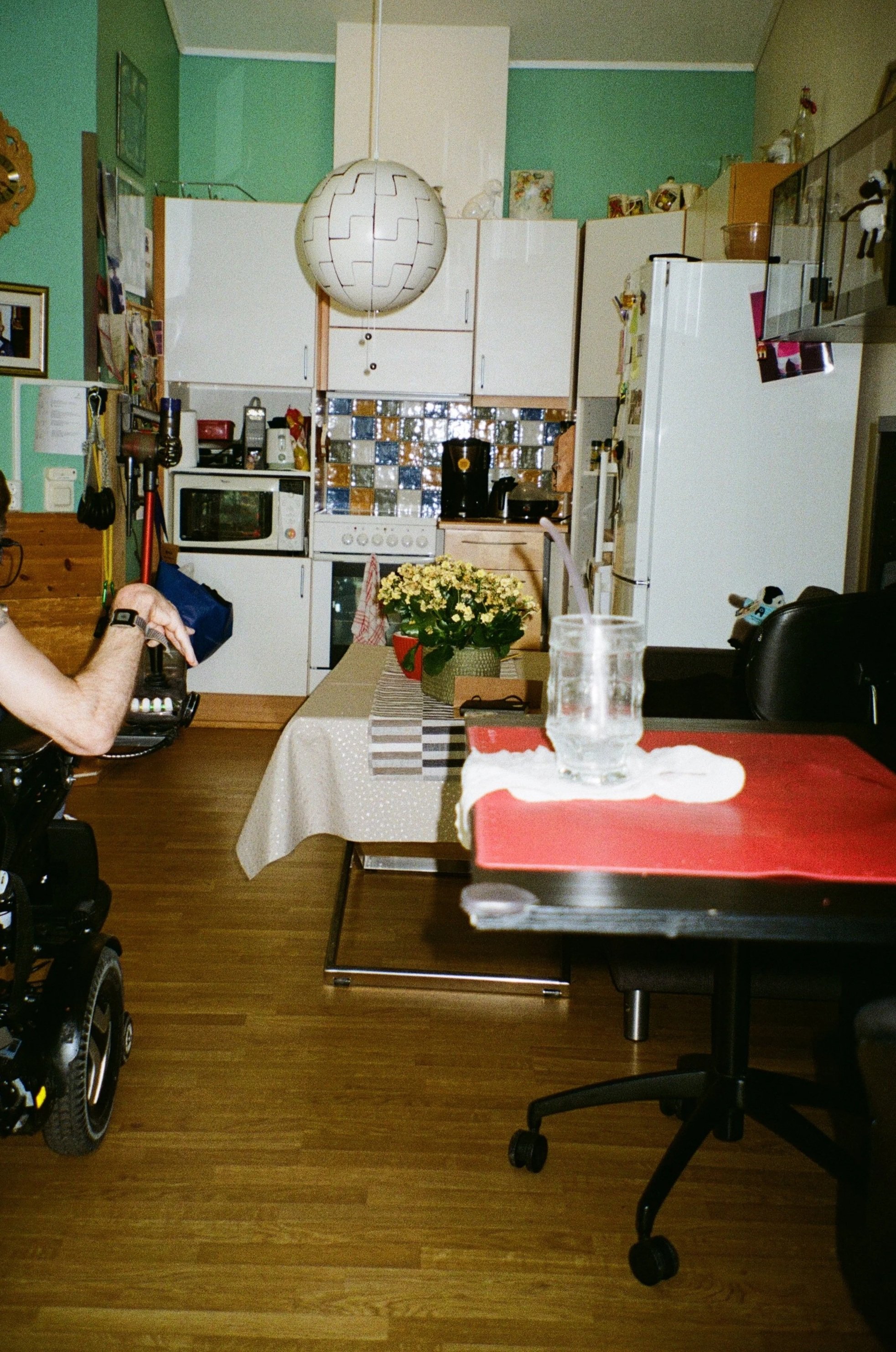 foto seende inn mot kjøkkenkrok. med en langt bord som stenger for åpningen og et lite bord foran det igjen. Skimter en person i en rullestol som står utenfor kroken og ser inn.