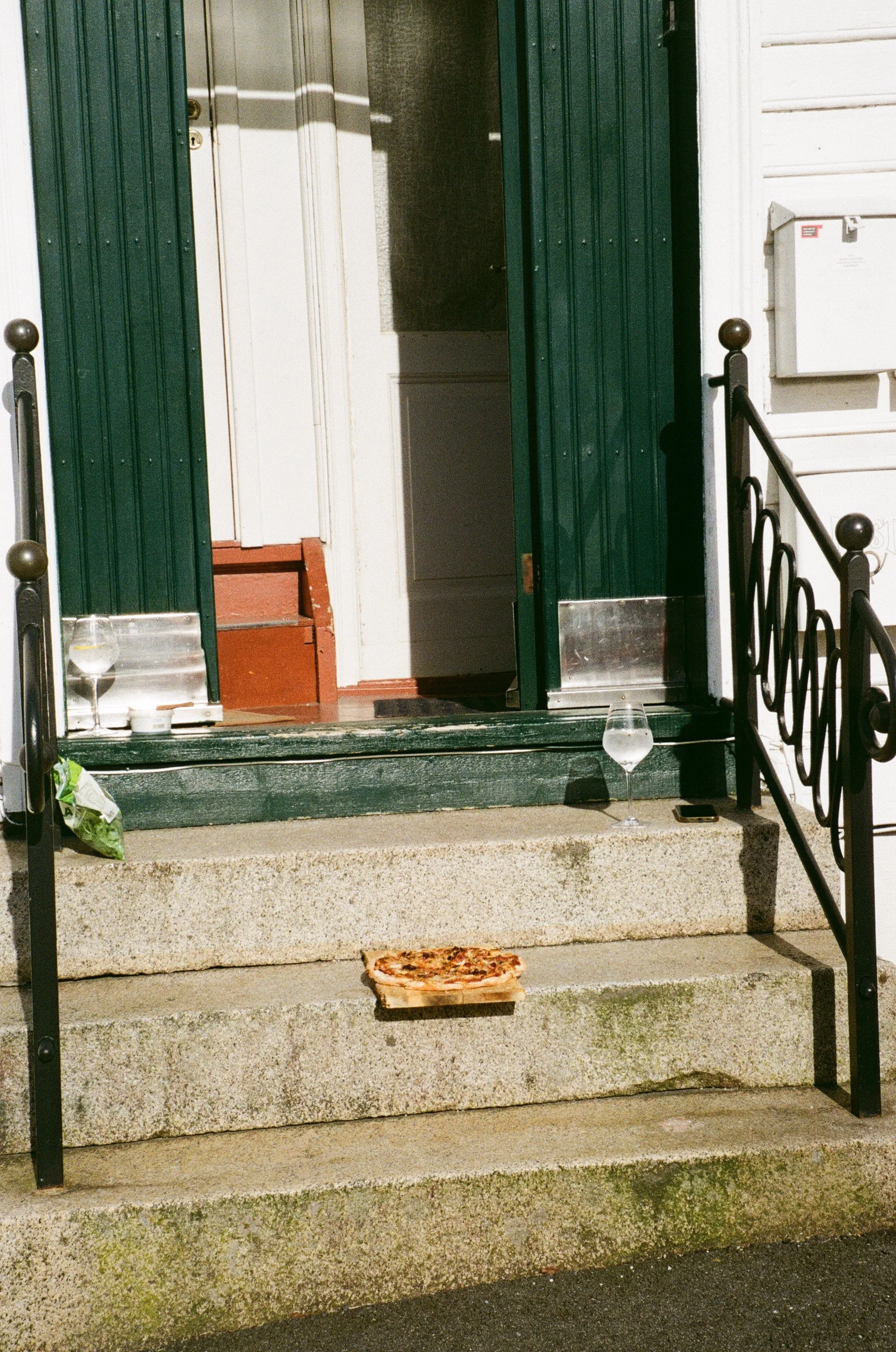 En del av en inngang til et hjem. døren står åpen. solen skinner på trappa fremfor døren og litt inn i rommet. på trappetrinnene står det hint av liv. et glass. en pizza.