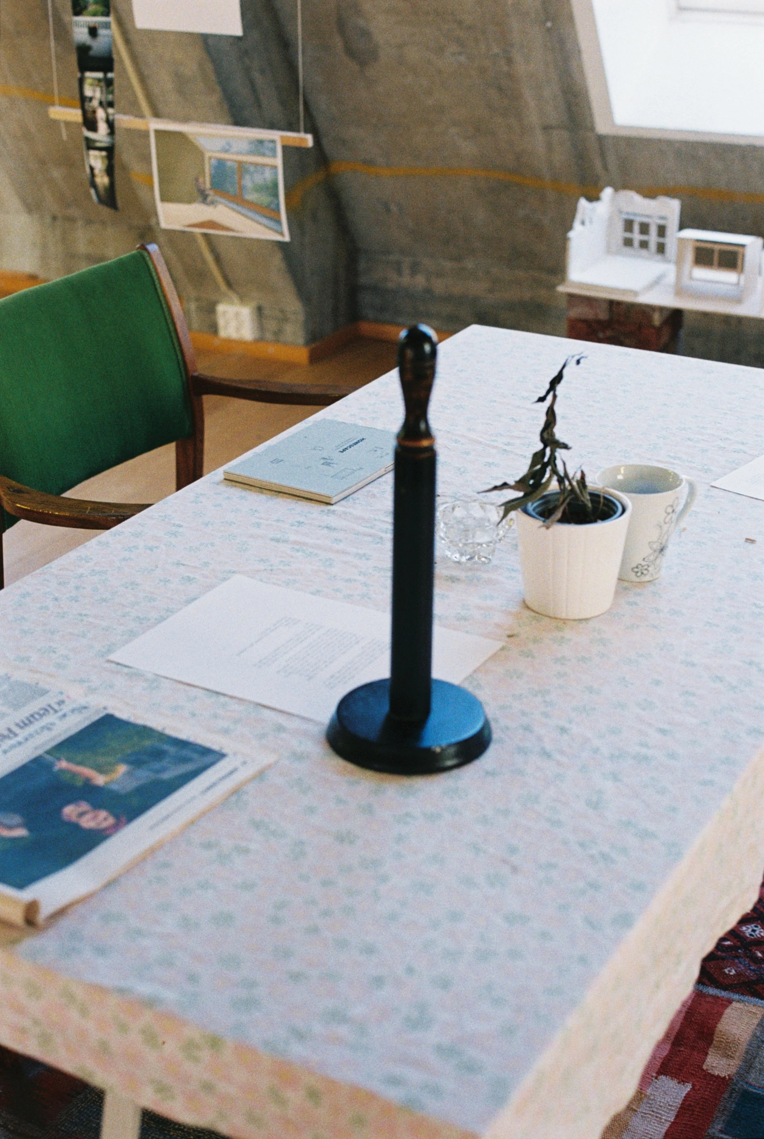 Foto fra utstilling. Kjøkkenbord med tørkerullholder, kaffekopp, liten plante, avis og en bok. kan så vidt se en stol inntil bordet.
