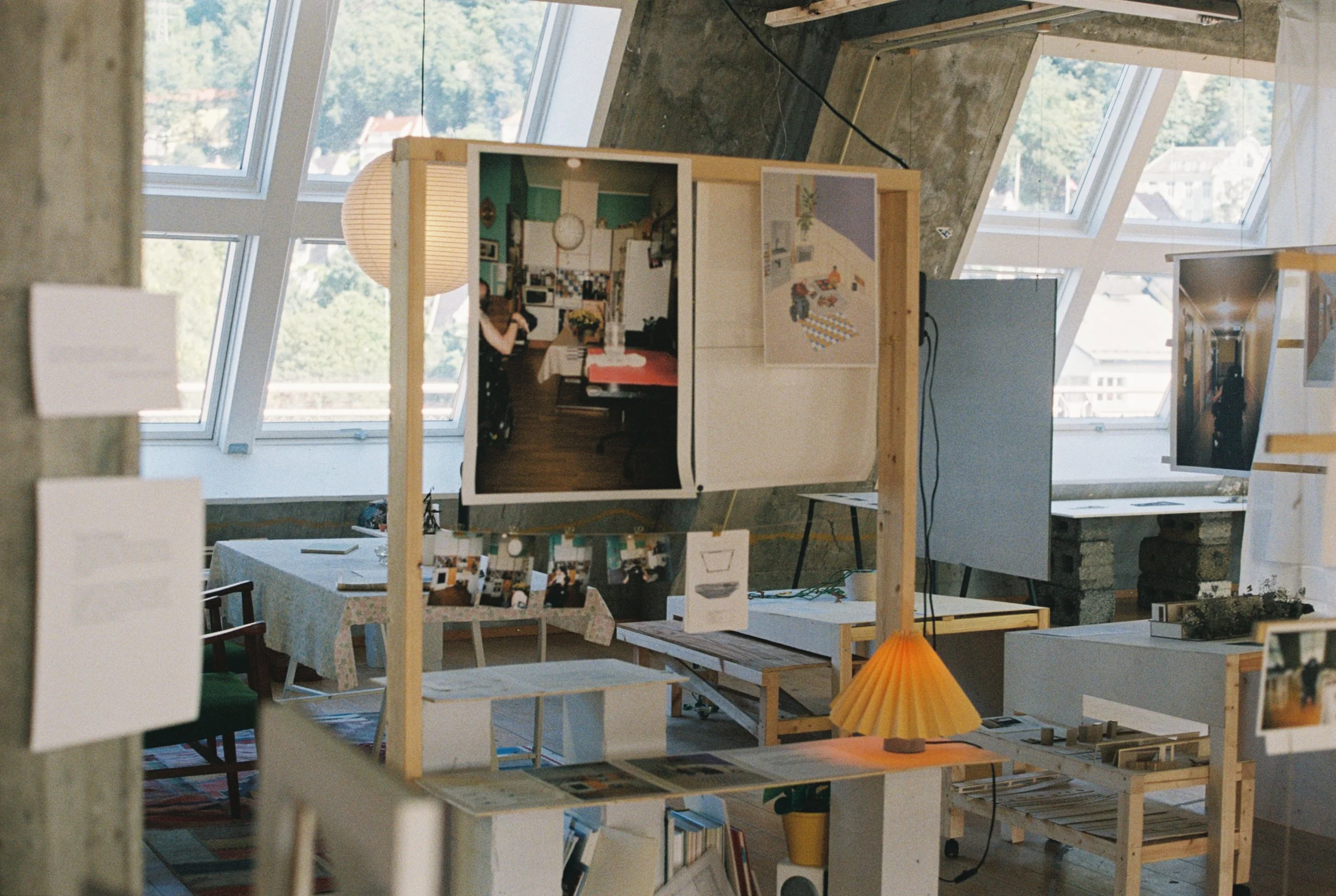 foto fra utstilling. åpent rom med stort vindu i bakgrunnen og stativ med bord, med foto og illustrasjoner på. skimter også et kjøkkenbordbord med stoler mellom stativ og vindu